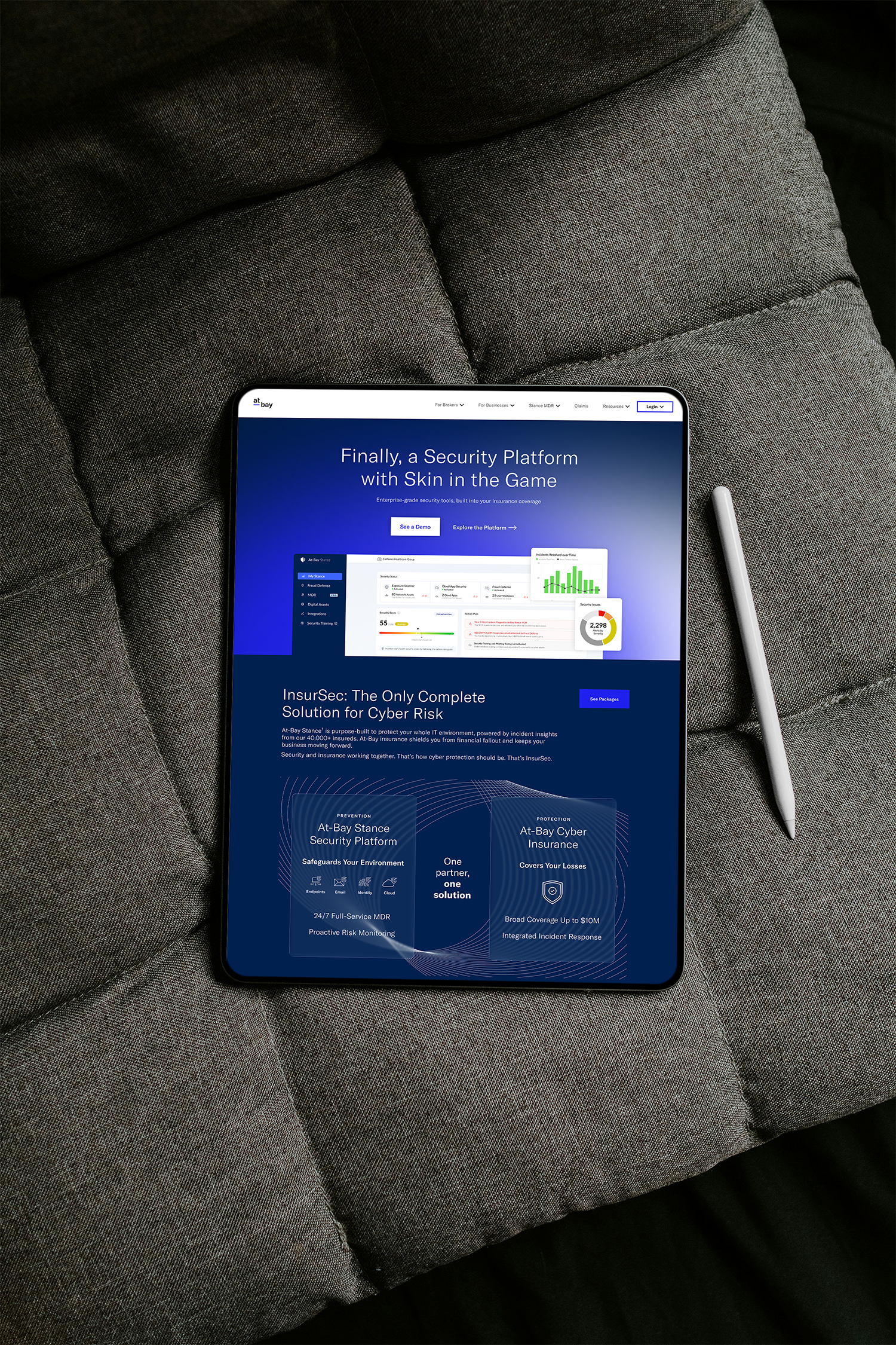 A tablet placed on a gray couch displaying a cybersecurity platform webpage with the headline "Finally, a Security Platform with Skin in the Game." Next to the tablet is a gray stylus.