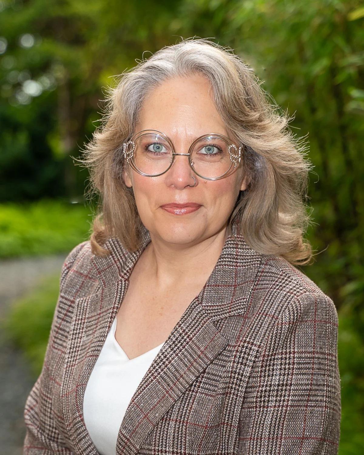 Tommie Lorene, wearing a plaid blazer over a white top, is outdoors with a blurred green foliage background.