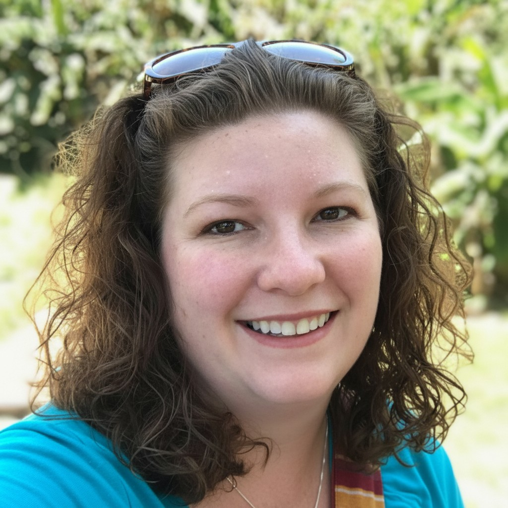 A woman with curly brown hair wearing sunglasses on her head and a blue top, smiling outdoors with greenery in the background.