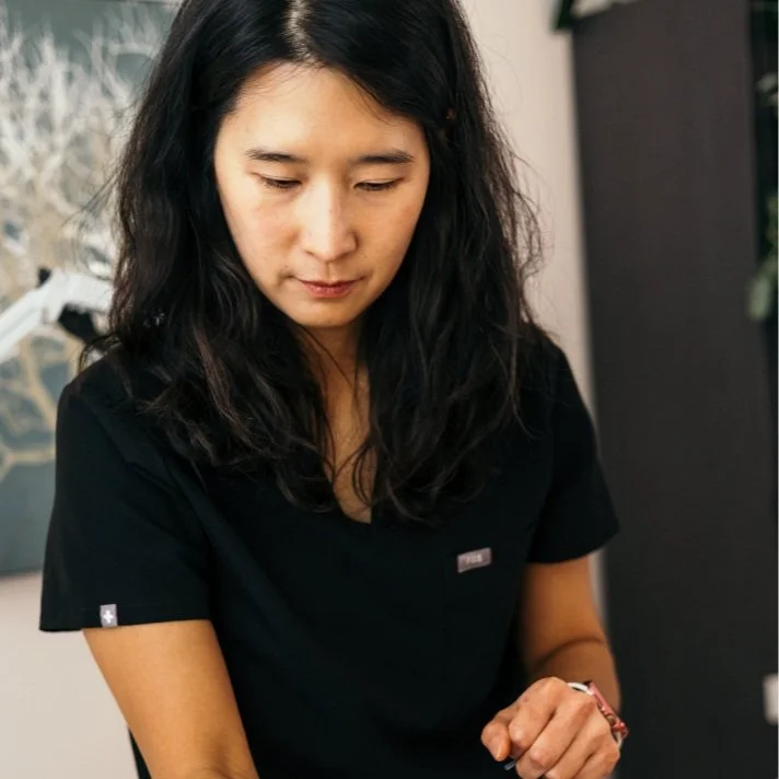 A woman with long black wavy hair wearing a black shirt is looking down, indoors with a blurred background.