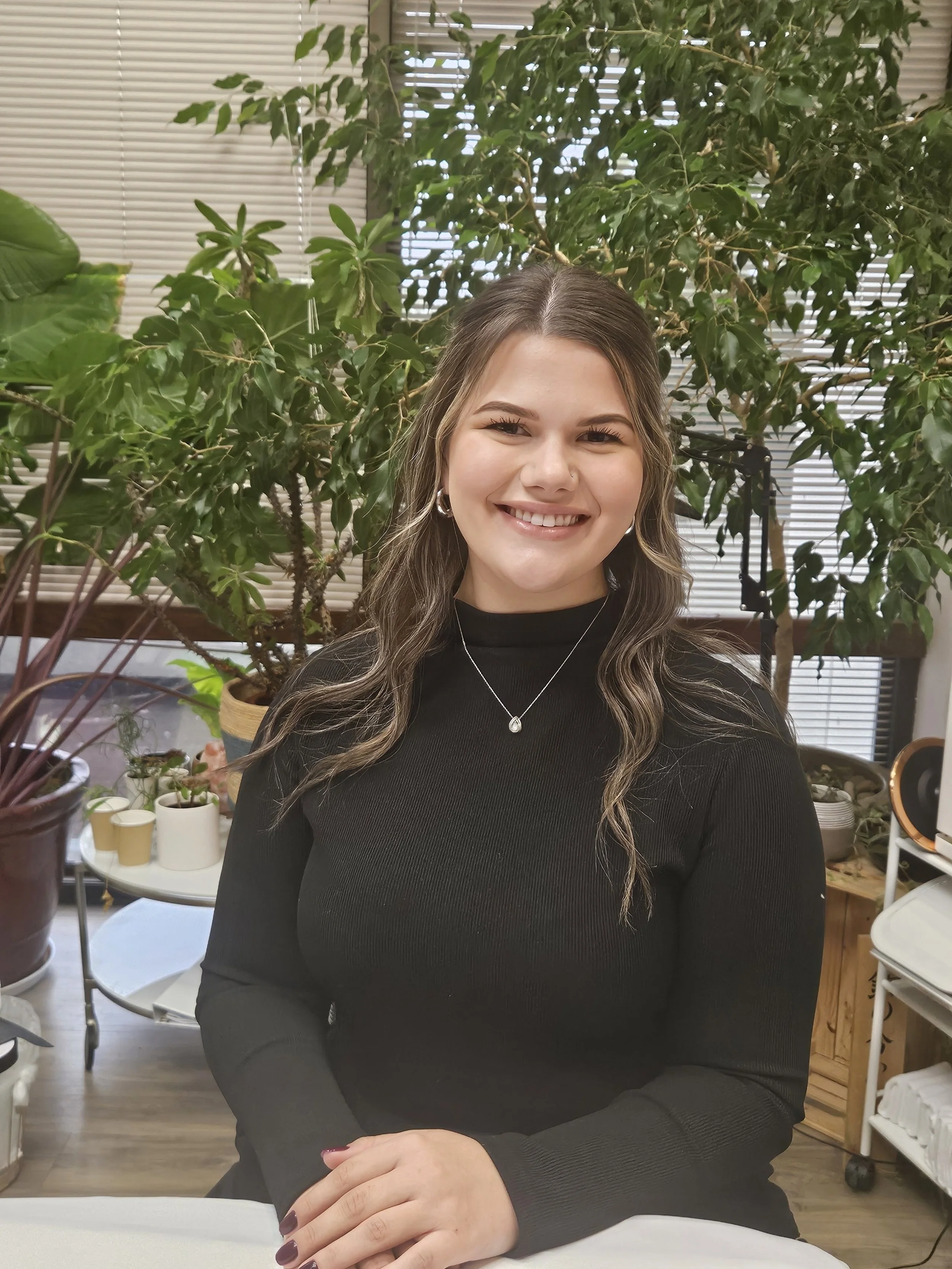 Smiling woman with long wavy hair wearing a black top, standing in front of lush green plants and window blinds.