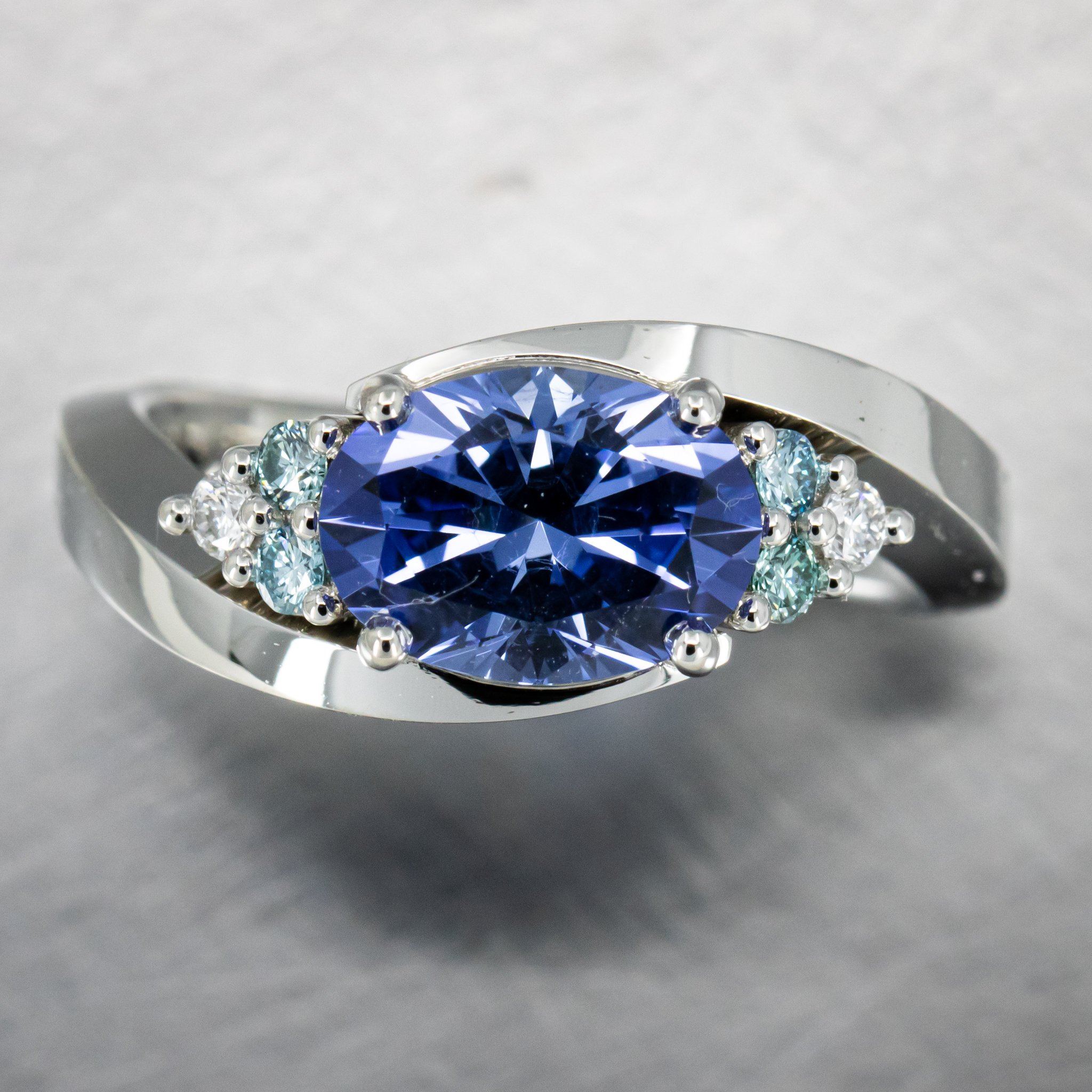 Custom Engagement Ring featuring a Laboratory Grown Oval Custom-Cut Cornflower Blue Laboratory Grown Sapphire with Two Round Brilliant Cut Irradiated Aqua Blue and One Colorless Diamond Situated on Either Side of the Center with an Assymmetrical Shan