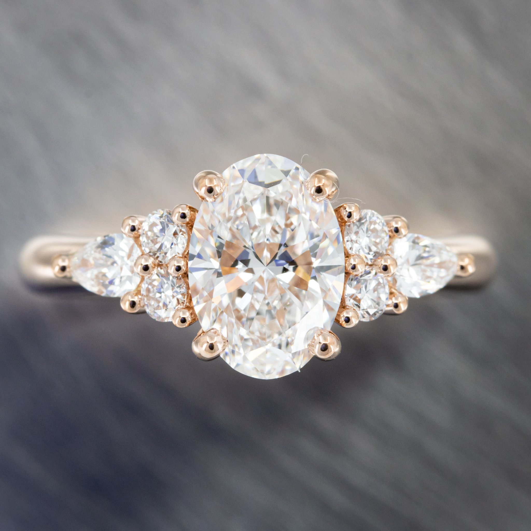 Custom Engagement Ring in 14k Rose Gold with an Oval Brilliant Cut Laboratory Grown Diamond in a Four Prong Setting with Two Round Brilliant Cut and One Pear Shape Laboratory Grown Diamond on Either Side of the Center Stone. Considering custom? Check