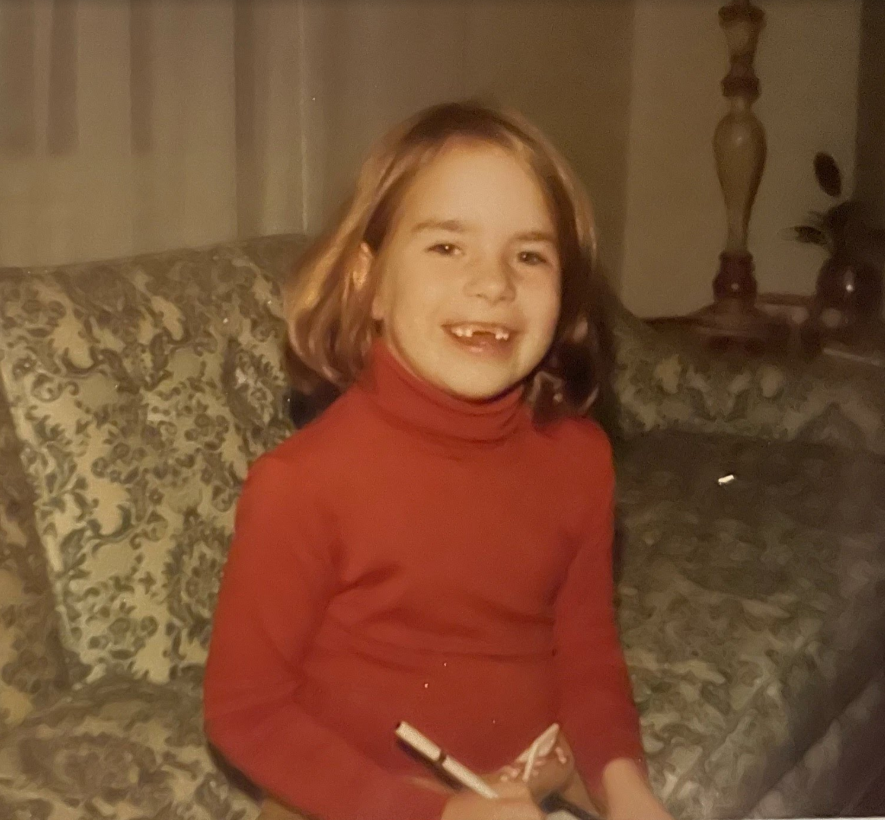 A young girl with short, light brown hair wearing a red turtleneck sweater, smiling and holding a pen, sitting on a patterned couch.