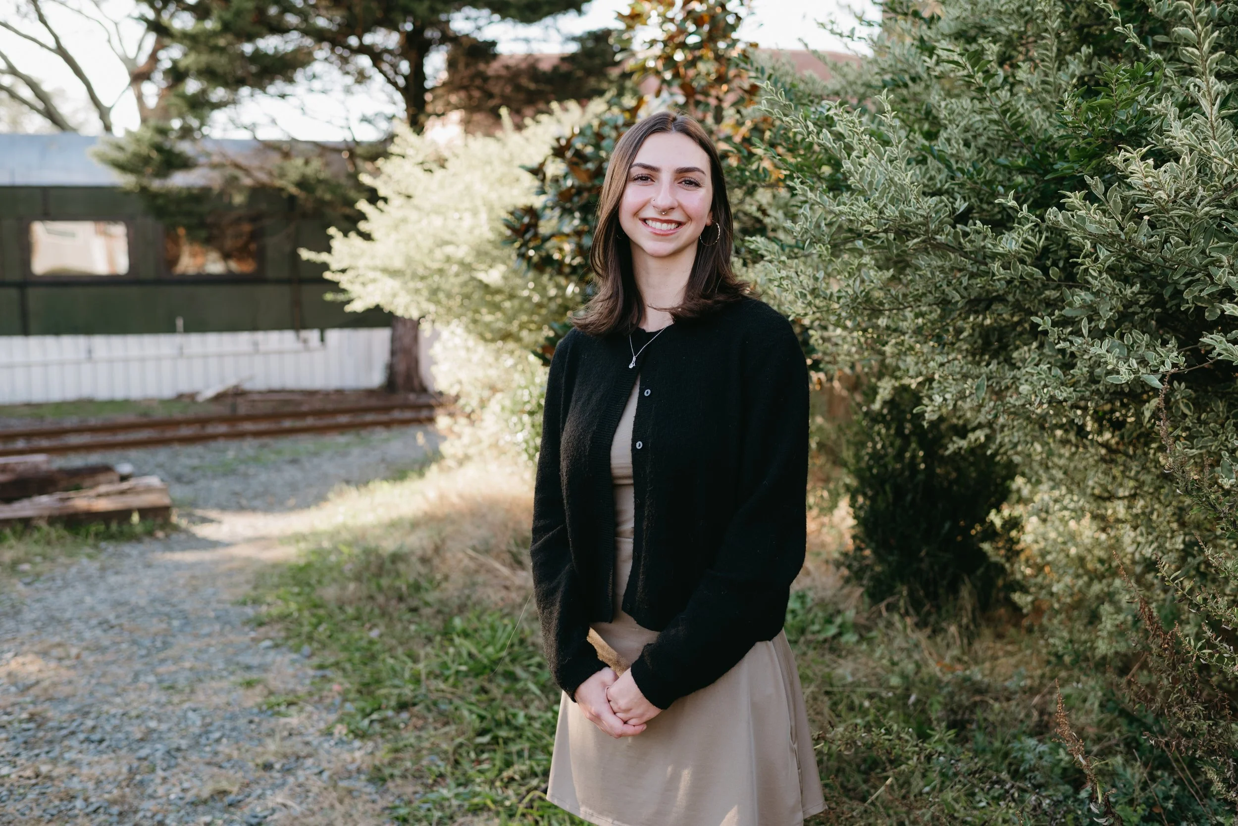 A young woman with shoulder-length dark hair, smiling, standing outdoors in front of green bushes and trees, wearing a black cardigan and beige dress.