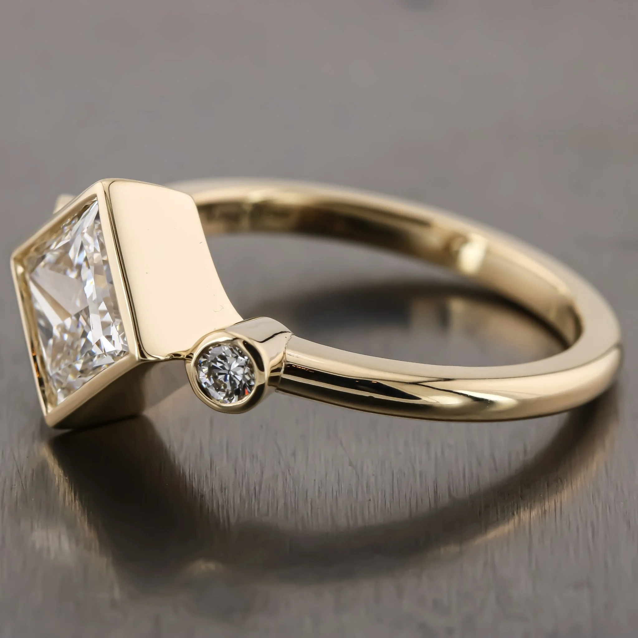 Close-up of a gold ring with a square-shaped diamond on top and a small round diamond on the side, resting on a reflective dark surface.