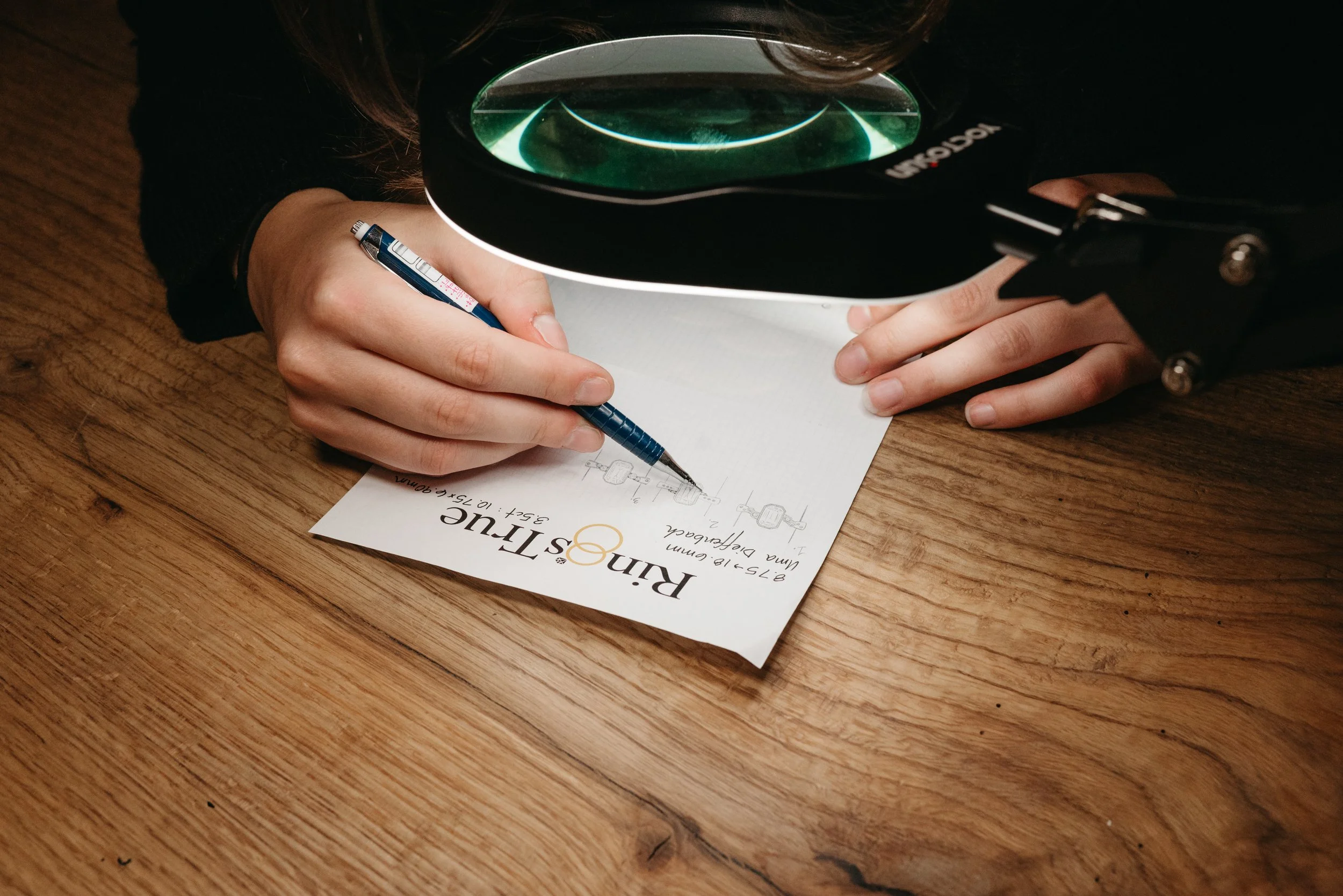Person using a magnifying glass while drawing technical sketches on a paper titled 'Rin-Stir' placed on a wooden table.