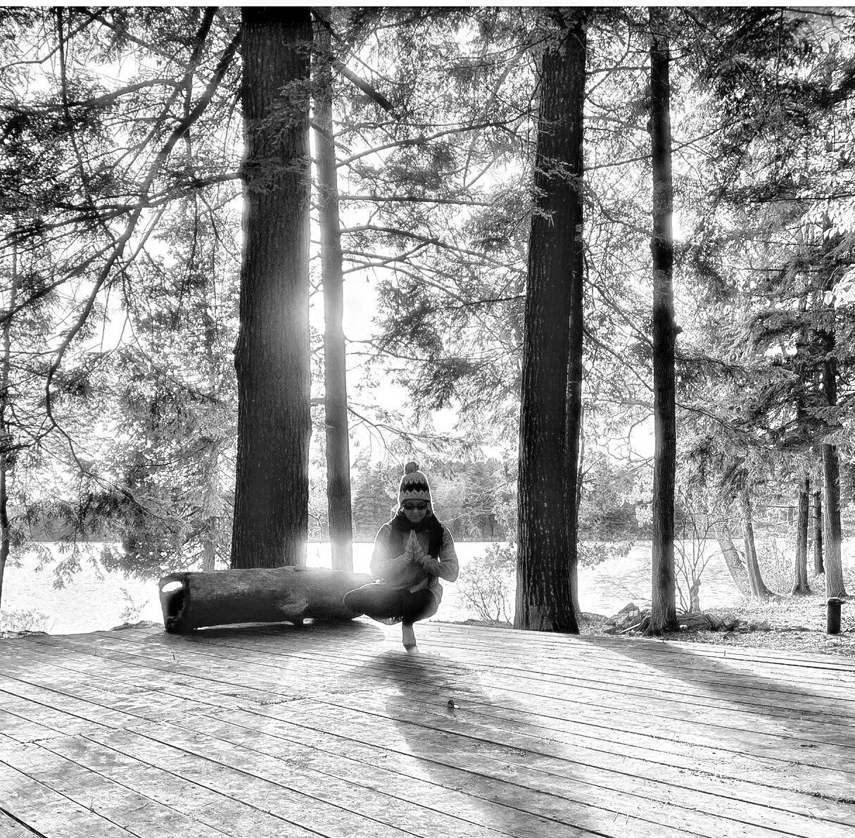 A person in a beanie and glasses practicing yoga or meditating on a wooden dock near a forested area with trees and water, backlit by sunlight.