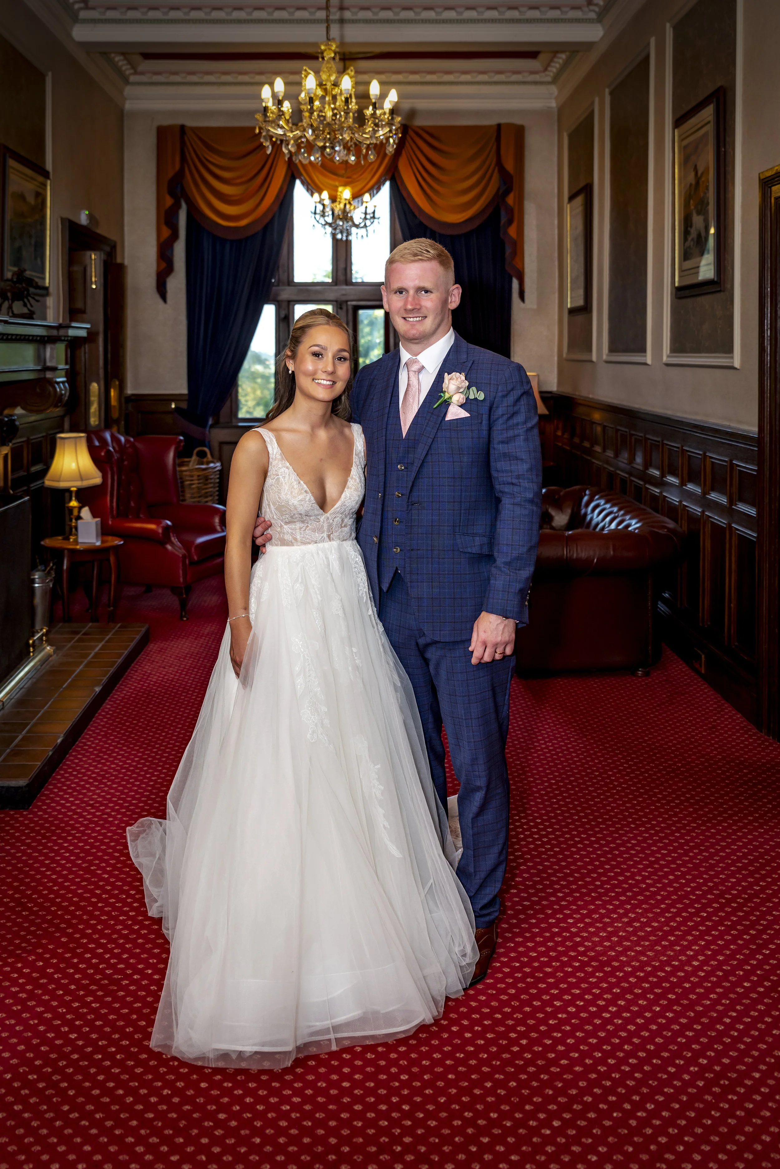 A bride and groom standing in a warmly decorated room with a staircase, red carpet, and elegant furnishings.