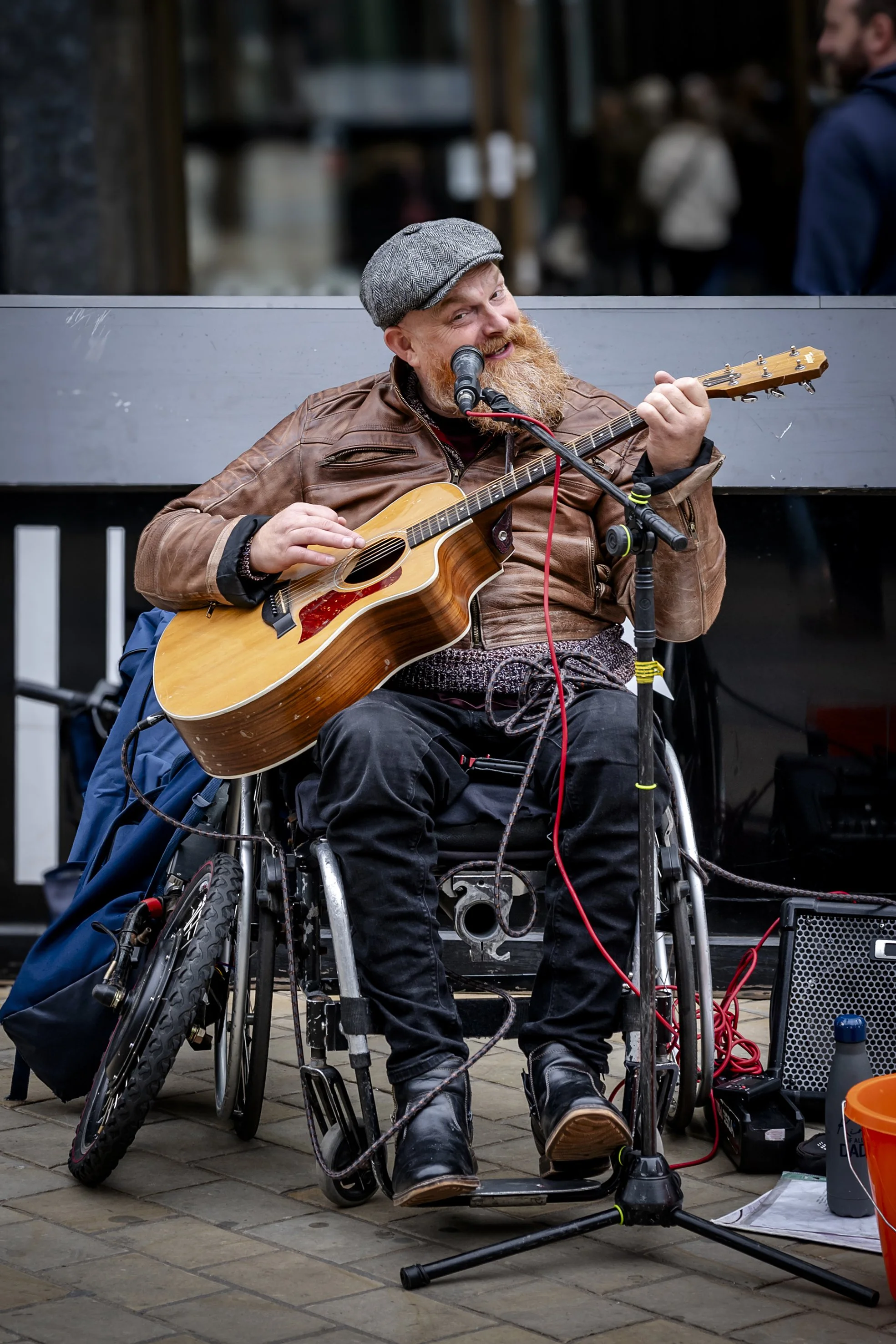 A man in a wheelchair playing an acoustic guitar and singing into a microphone on a street, smiling and wearing a brown leather jacket and gray flat cap.