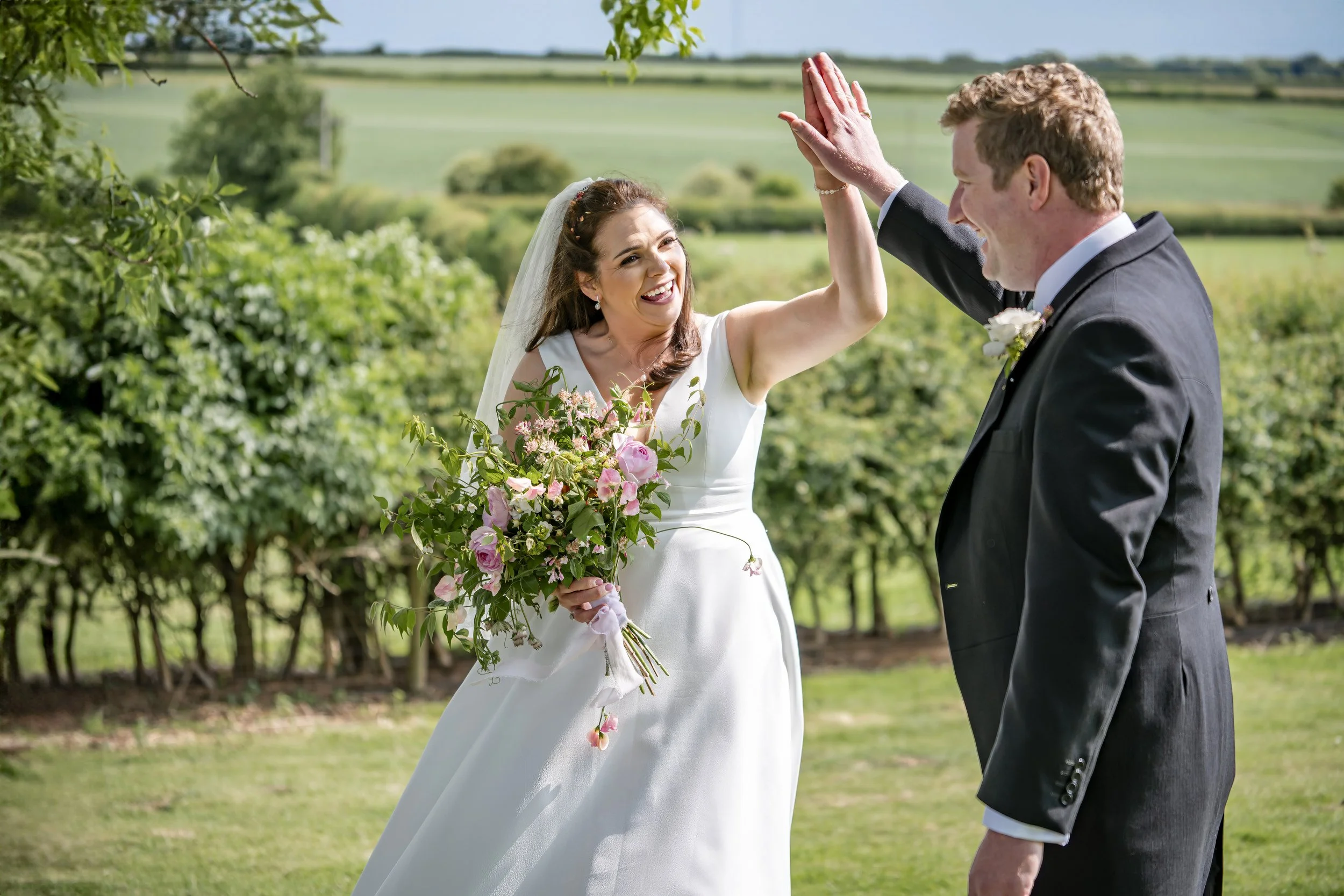 A bride and groom high-fiving outdoors on a wedding day, with green fields and trees in the background.