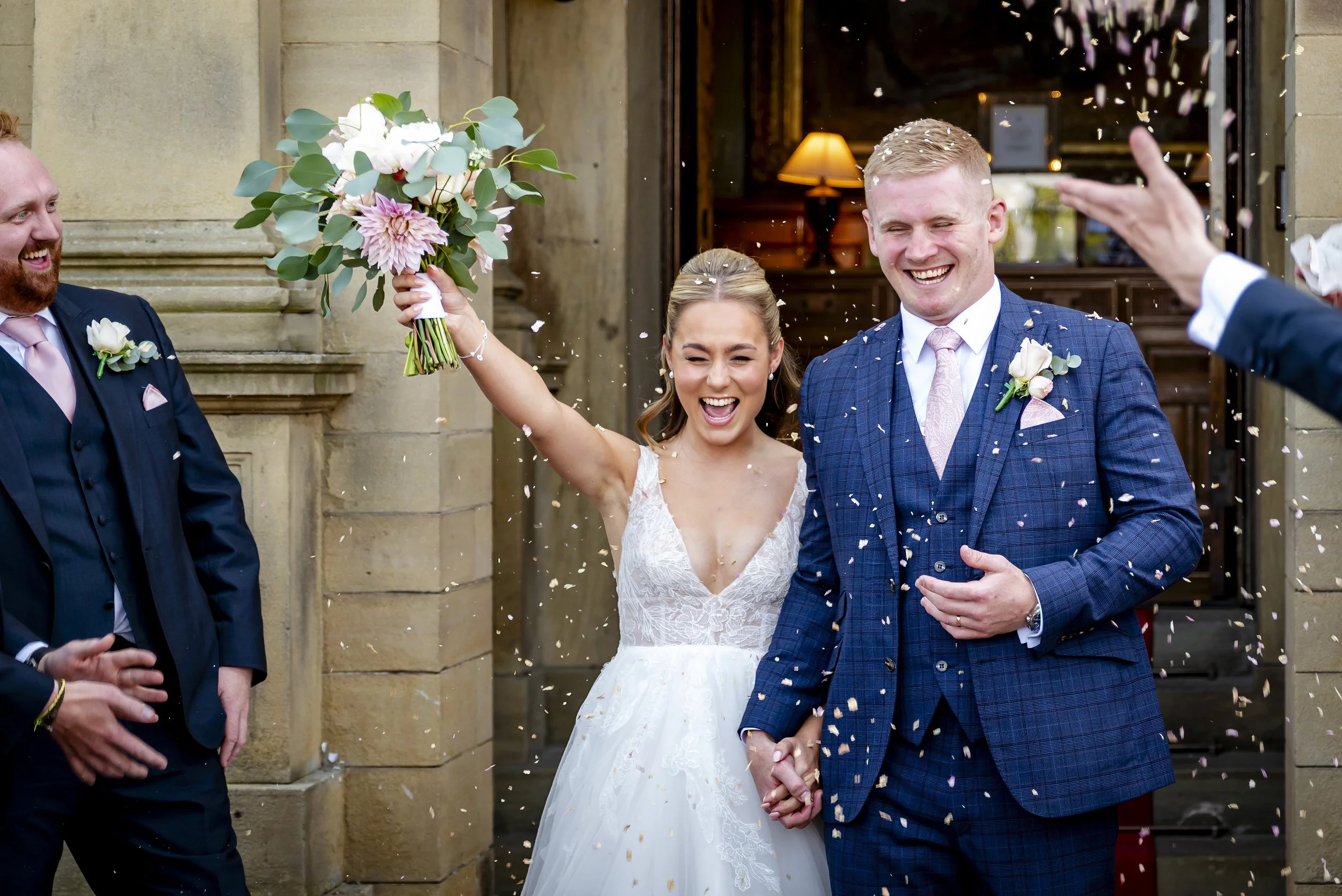 A newlywed couple holds hands and is celebrated outside a church, with guests throwing confetti. The bride is holding a bouquet of pink and white flowers. Both are smiling happily.