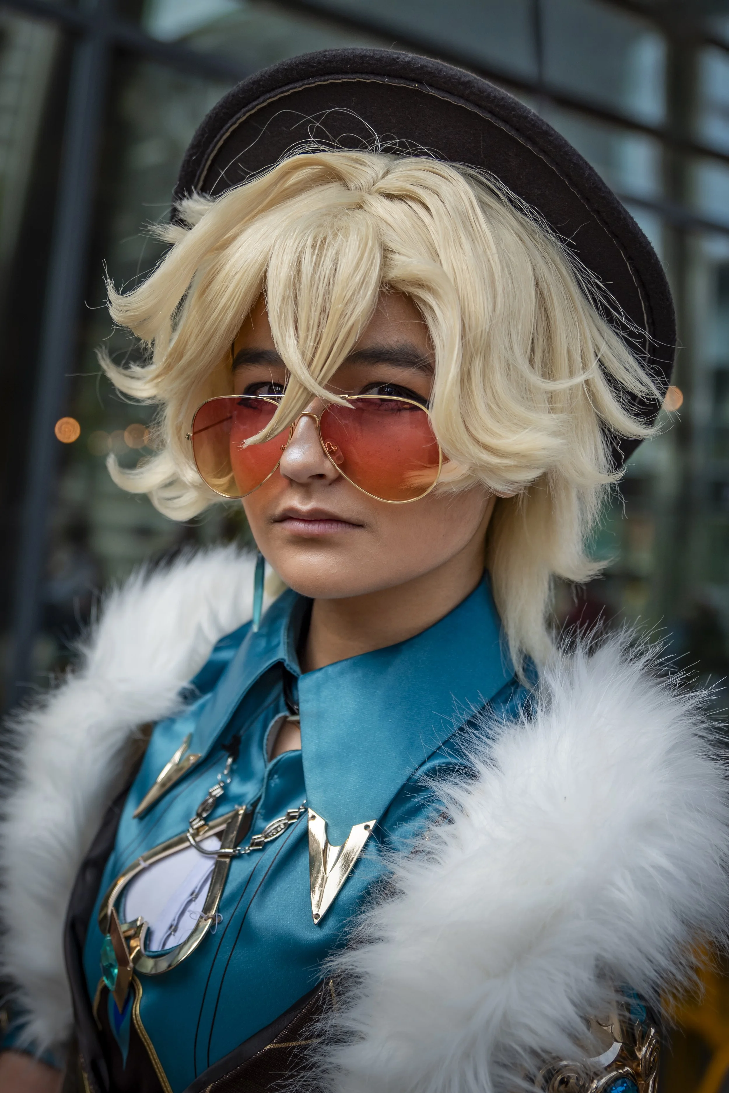 A person with platinum blonde, wavy hair wearing pink-tinted aviator sunglasses, a black hat, and a blue outfit with metallic accessories, and a white furry coat.