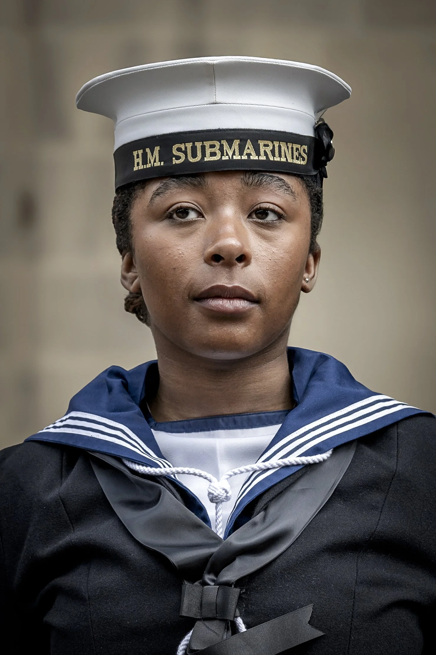 A young woman in a navy and white Navy graduation cap and gown, with a serious expression, standing indoors.
