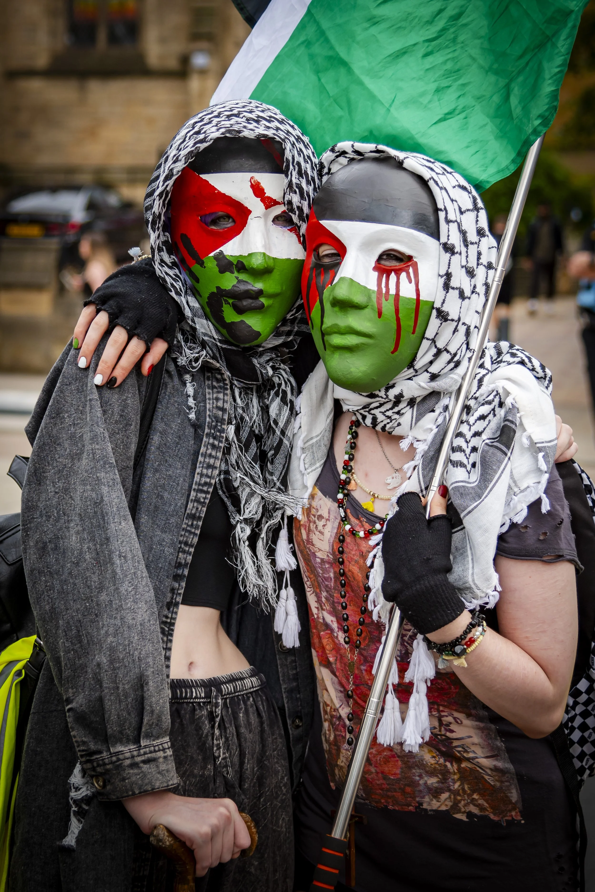 Two people wearing masks with Palestinian flag colors, embracing each other during a public event, holding an Iraqi flag. The masks have eye holes and depict crying and blood-dripping effects. One person is wearing a black jacket with a patterned hea