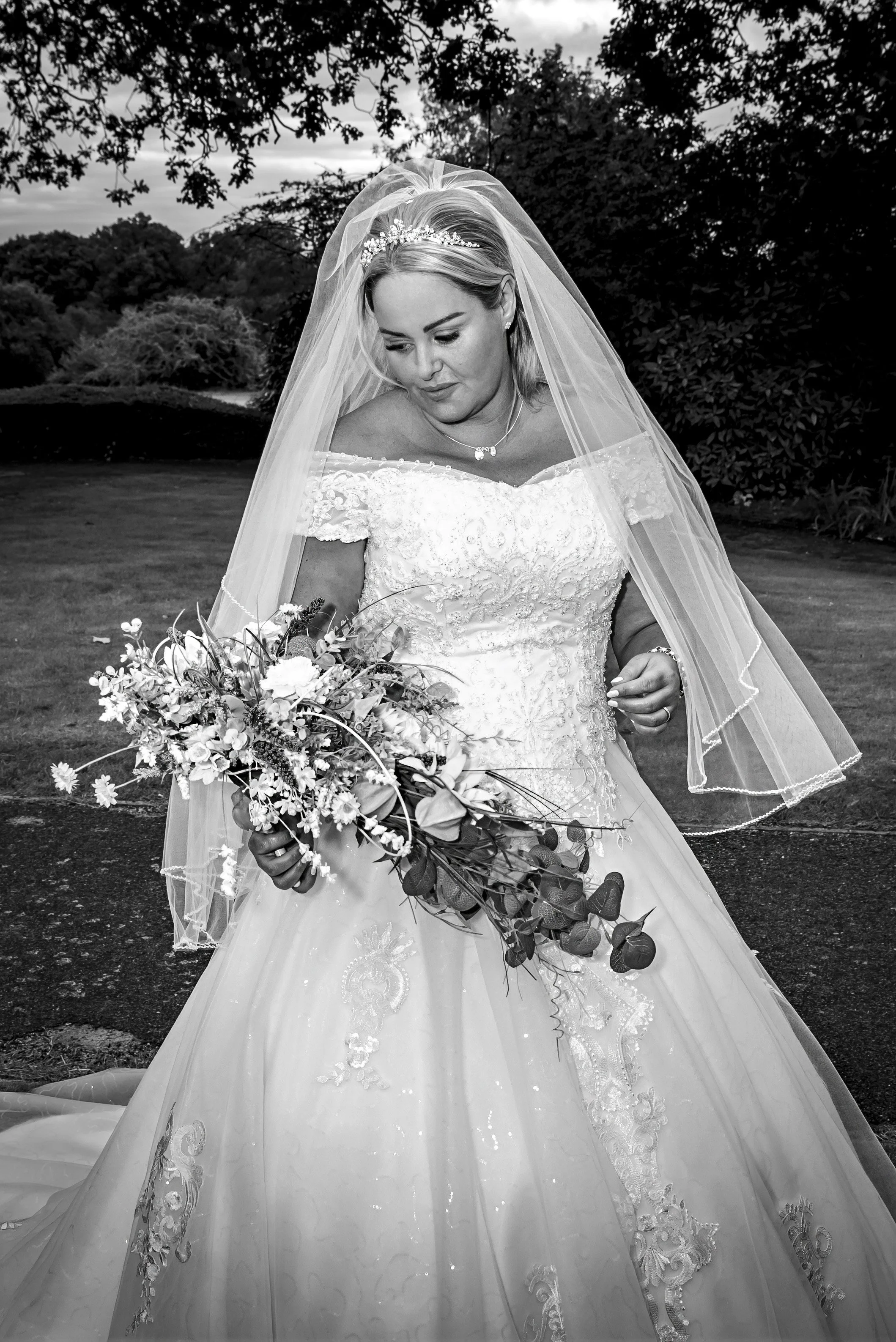 Black and white photo of a bride in a wedding dress holding a bouquet of flowers, outdoors with trees in the background.