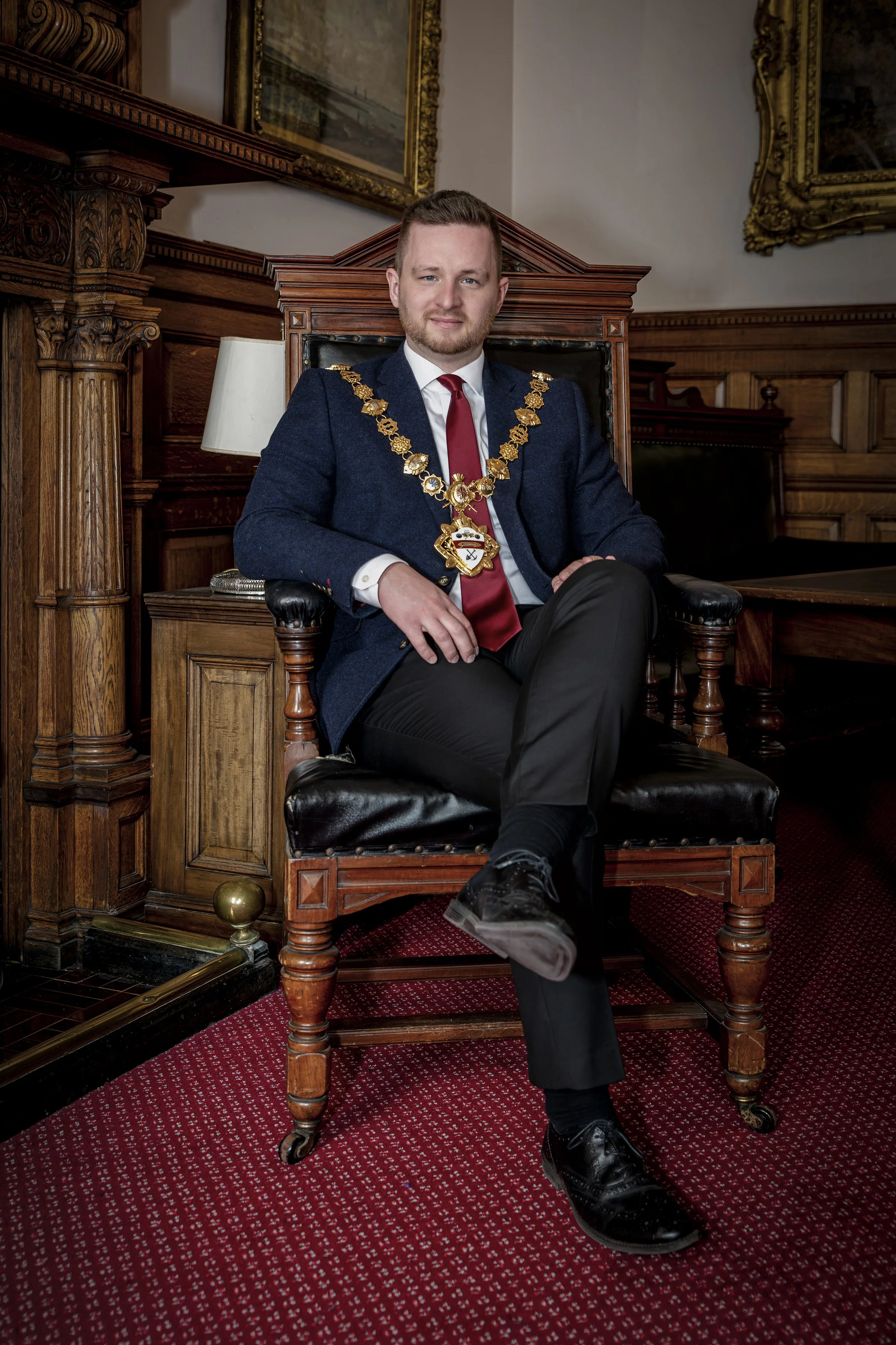 A young man in a suit sitting on a wooden chair with a green and gold chain of office around his neck in a wood-paneled room with framed pictures on the walls.