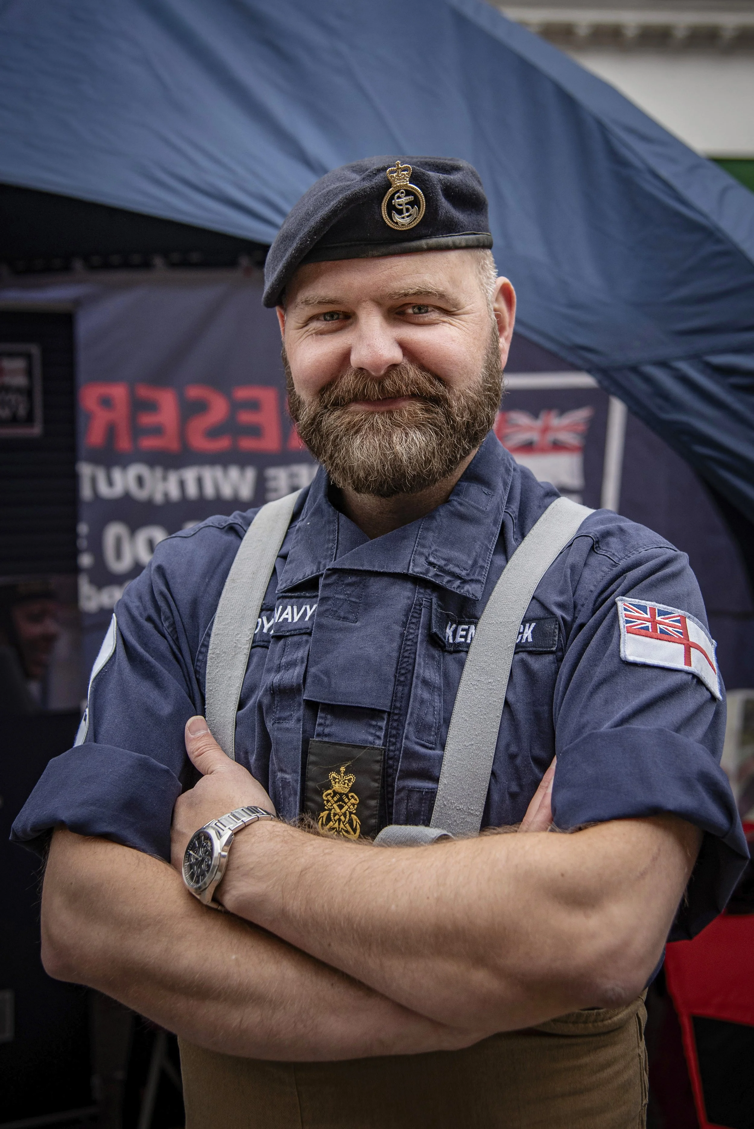 A man in a navy uniform with British flags on his sleeve and beret, standing with arms crossed and smiling in front of a blue tent.
