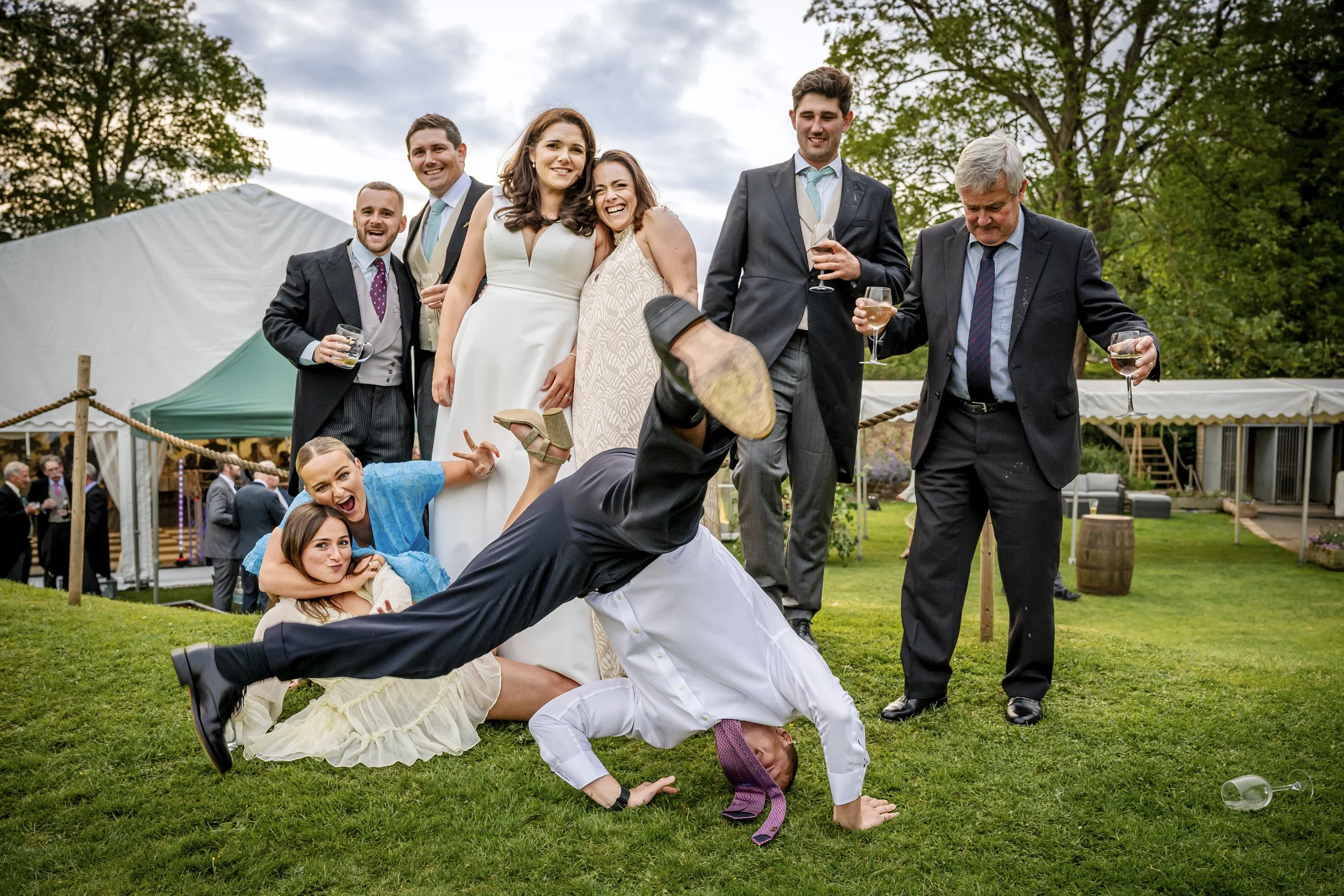 Group of people at a wedding or celebration outdoors, with some standing and some on the ground, engaging in playful activities, holding drinks, and smiling. A tent and trees are in the background.