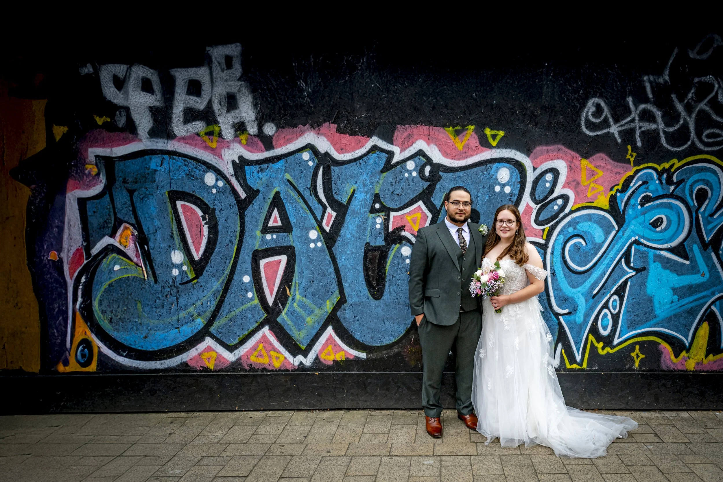 A newlywed couple stands in front of a colorful graffiti wall. The groom is wearing a dark suit, white shirt, and tie, and the bride is wearing a white wedding gown, holding a bouquet of flowers.