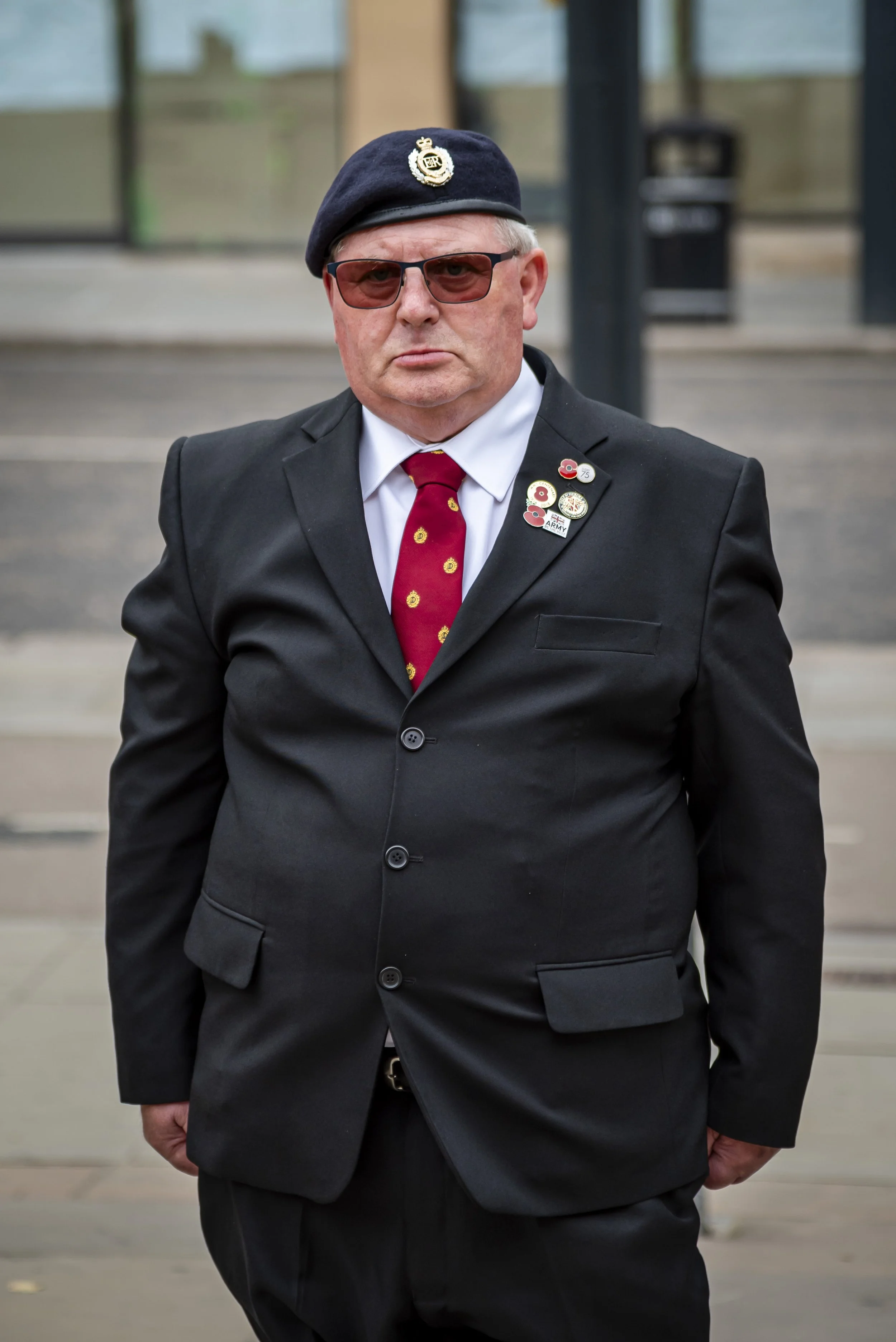 Older man in a black suit, white shirt, red tie with yellow designs, and a navy beret with badges. He also wears sunglasses, standing outdoors.