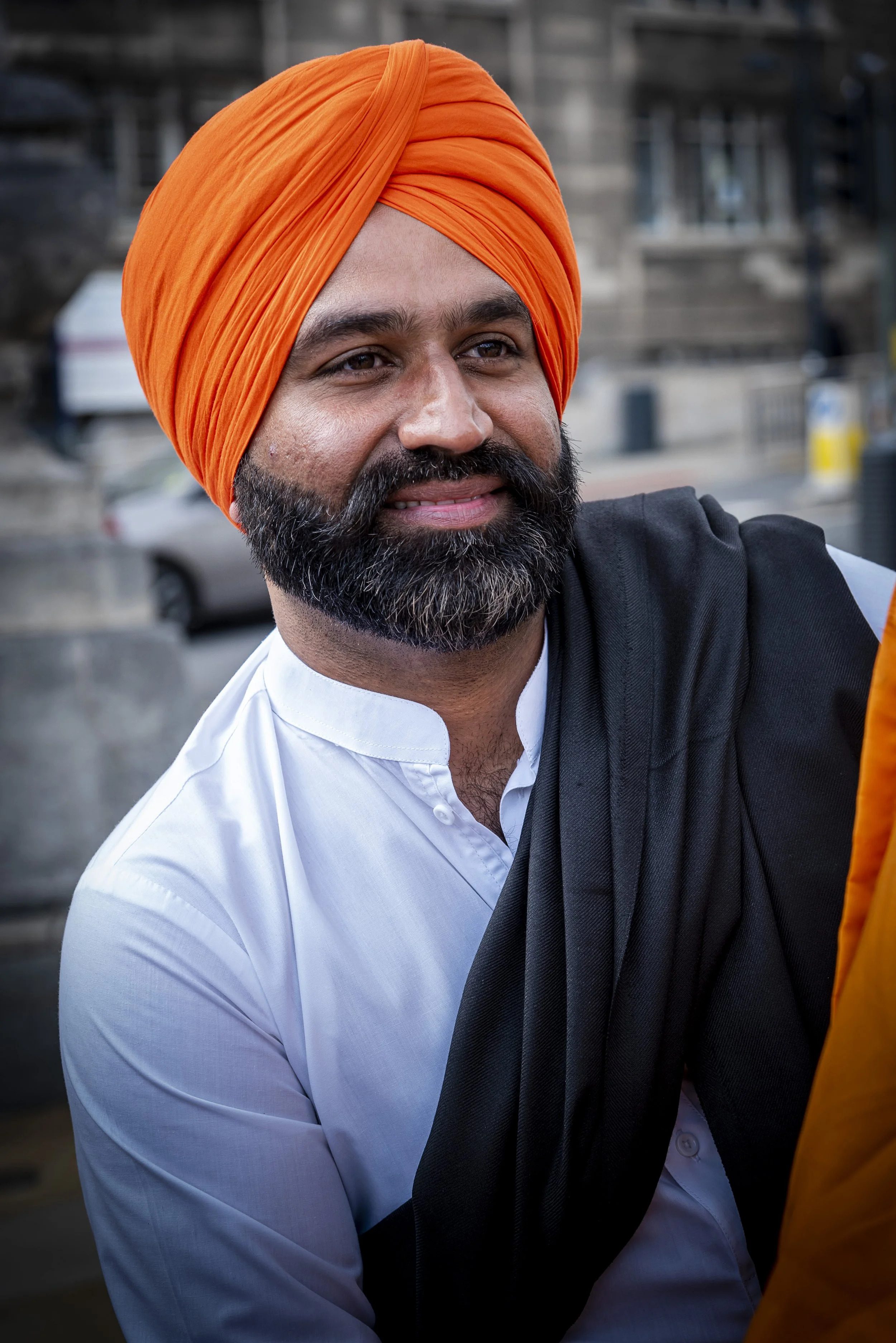 A man wearing an orange turban, white shirt, and black shawl, smiling outdoors. There are cars and urban buildings in the background.