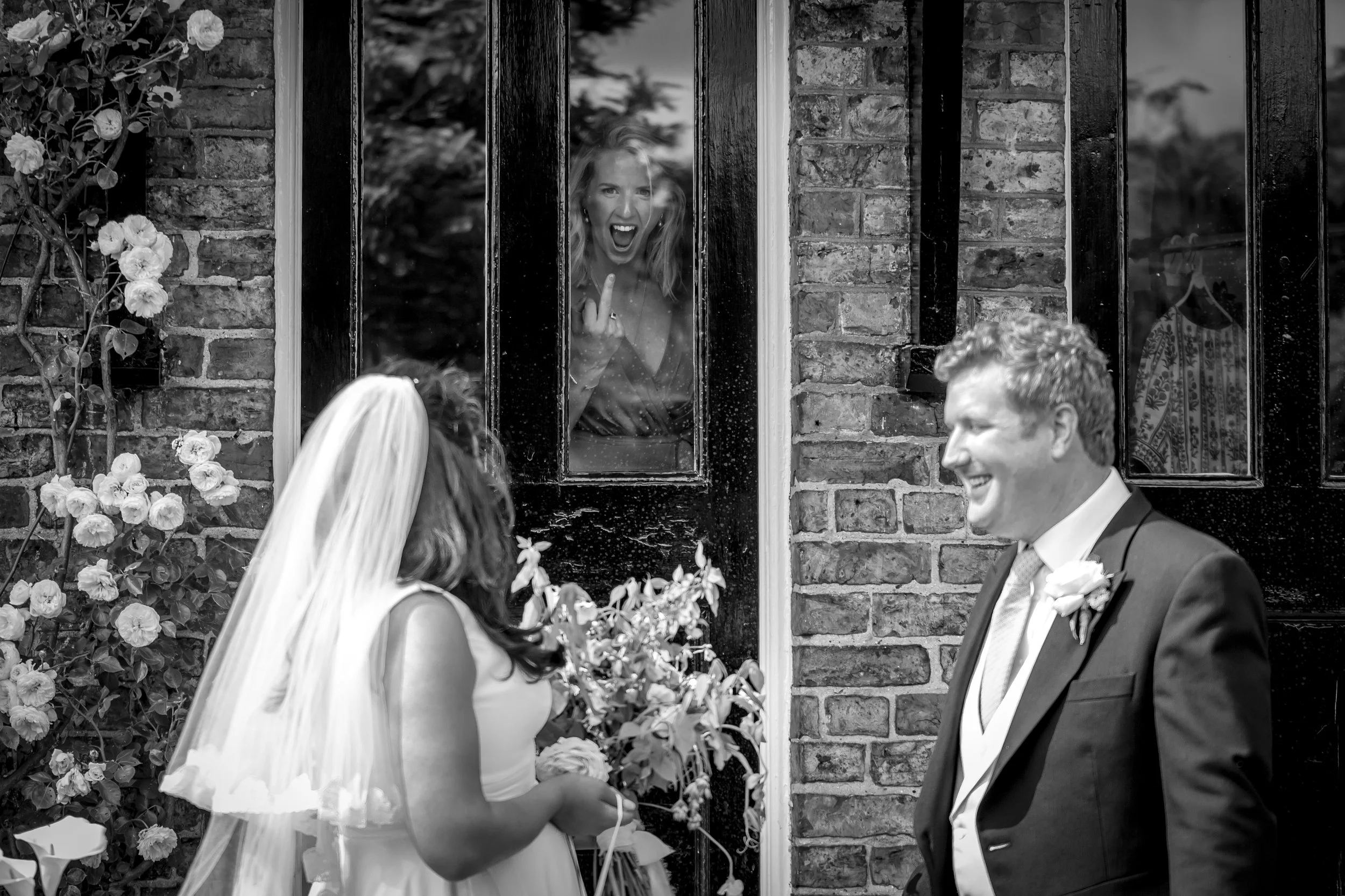 A bride and groom are standing outside in front of a brick building. A woman inside, visible through a window, is smiling and making a rude gesture with her middle finger.