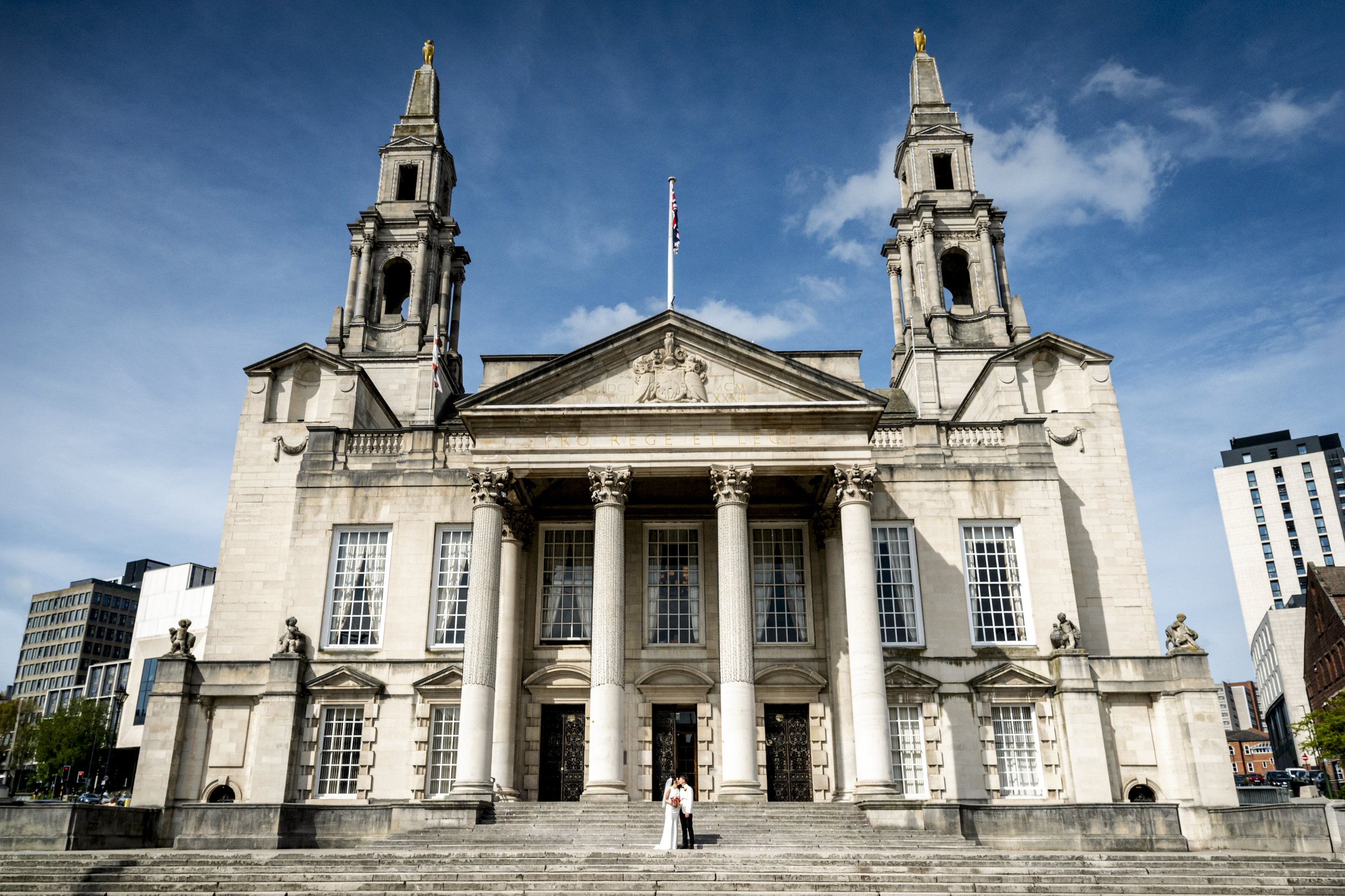 The large stone church with two tall towers in a city setting. The church has columns at the entrance, a coat of arms, and statues on the facade. A couple is standing in front of the church, with steps leading up to the entrance. Modern buildings are