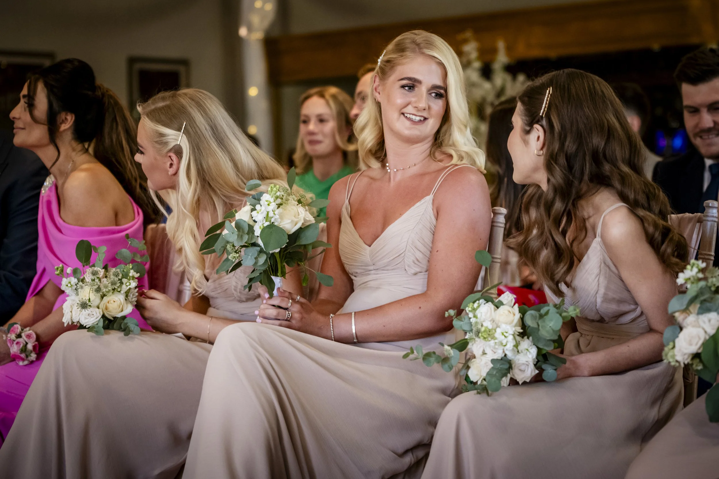 A group of women sitting in a row during a wedding ceremony, holding bouquets of white flowers and greenery, dressed in elegant dresses, and engaging in conversation.