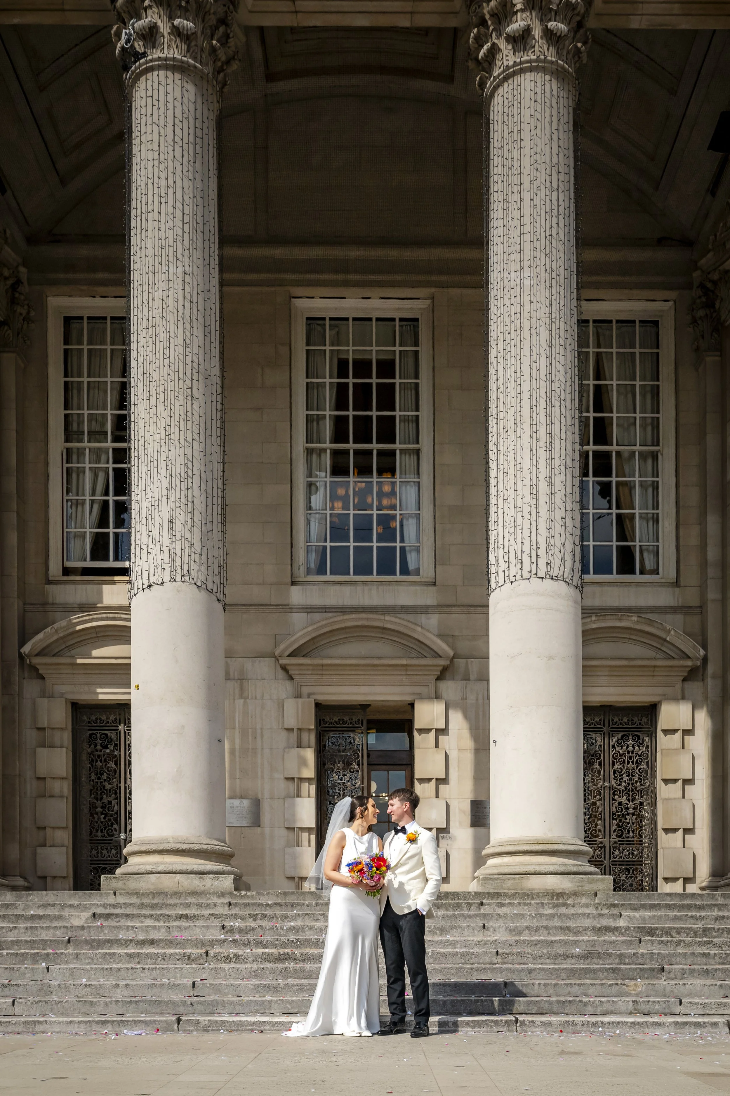 A bride and groom standing on steps outside a historic building with large columns. The bride is in a white wedding dress holding a bouquet, and the groom is in a white suit jacket, black bow tie, and black pants. They are smiling and looking at each