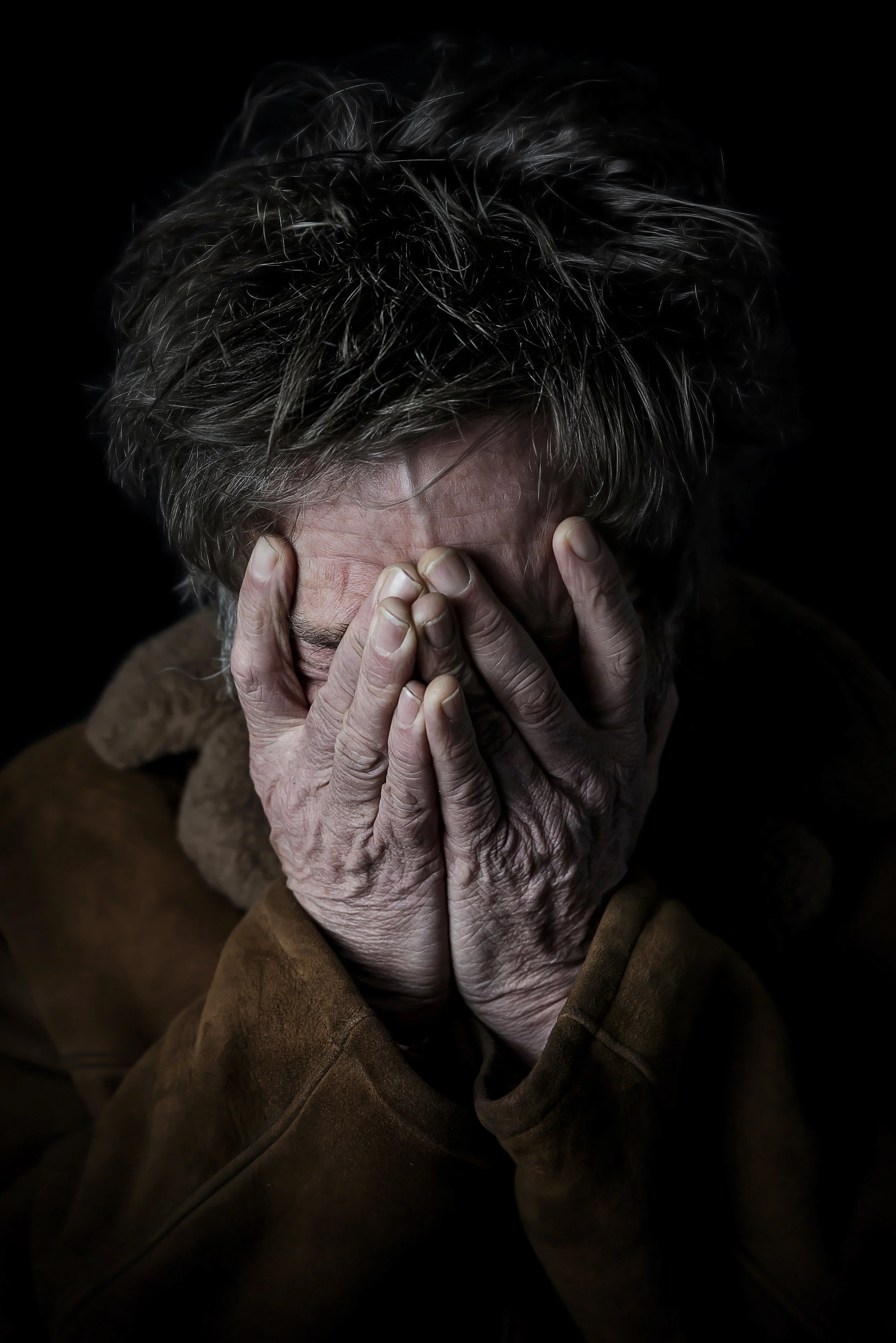 An elderly person with gray hair covering their face with both hands, wearing a brown jacket, against a black background.
