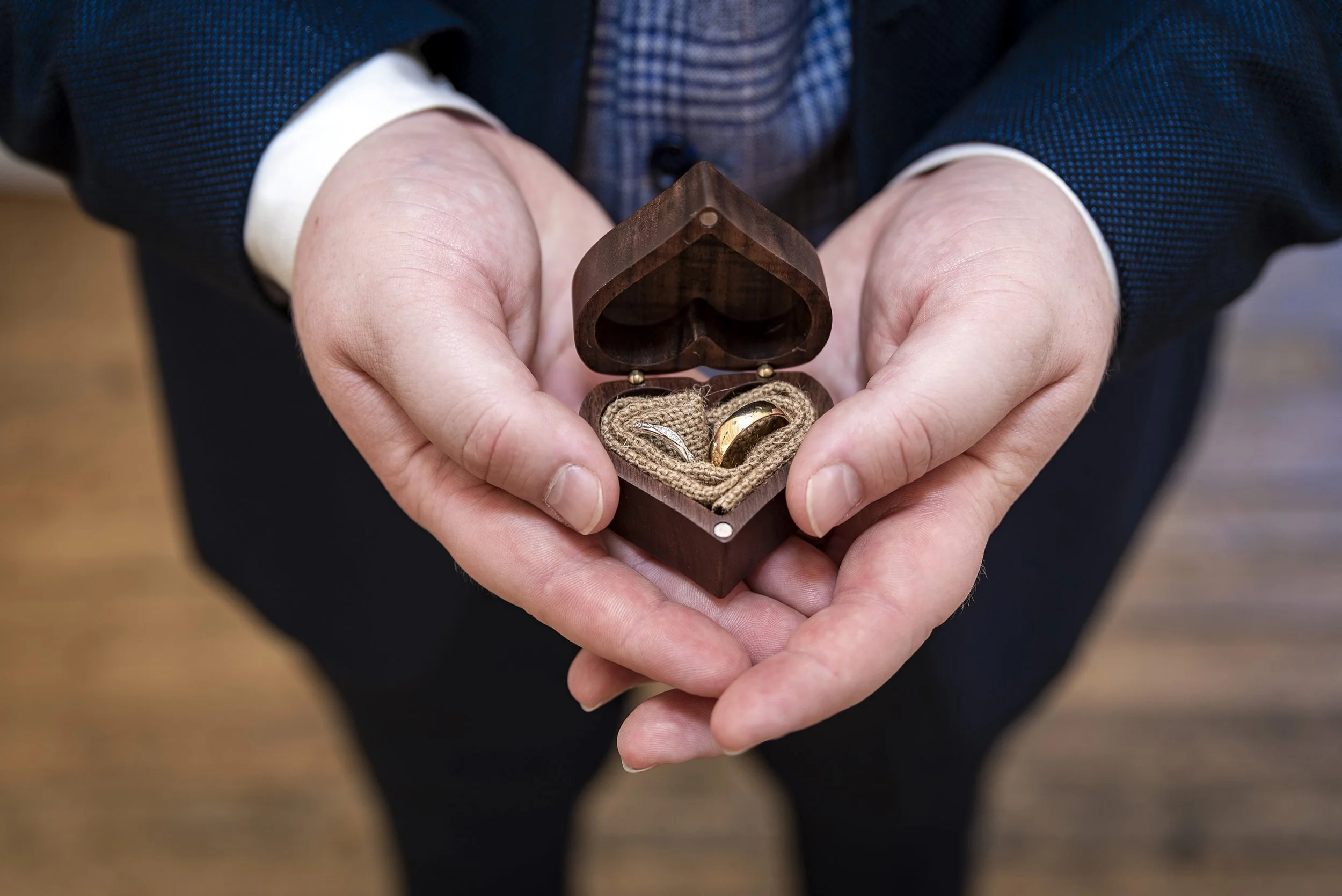 Person holding a dark wooden heart-shaped box containing two rings with a burlap cloth inside.