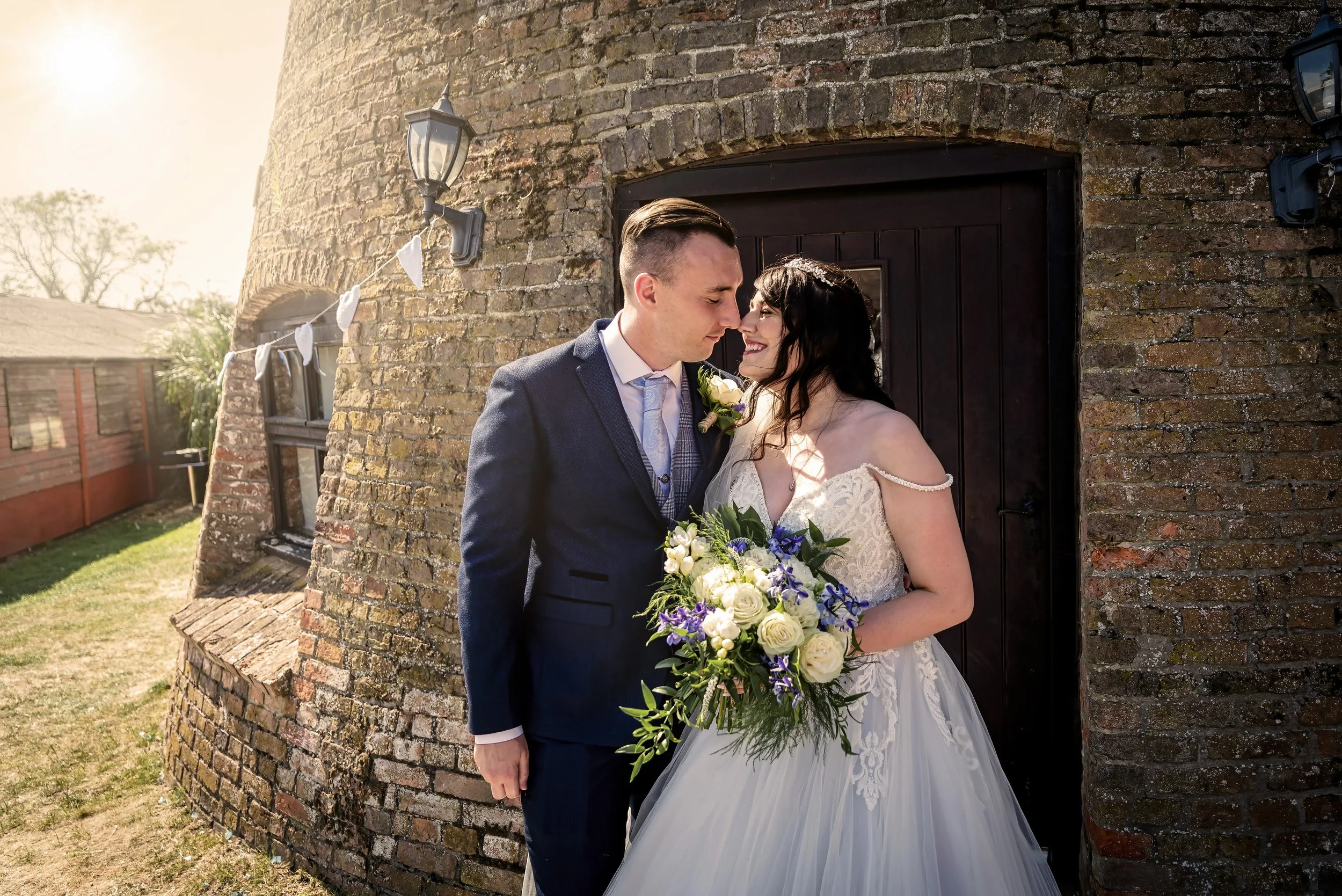 A bride and groom standing close together with their foreheads touching, smiling, in front of an old brick building with a small window and a lantern. The bride is holding a bouquet of white and purple flowers, and the groom is dressed in a dark suit