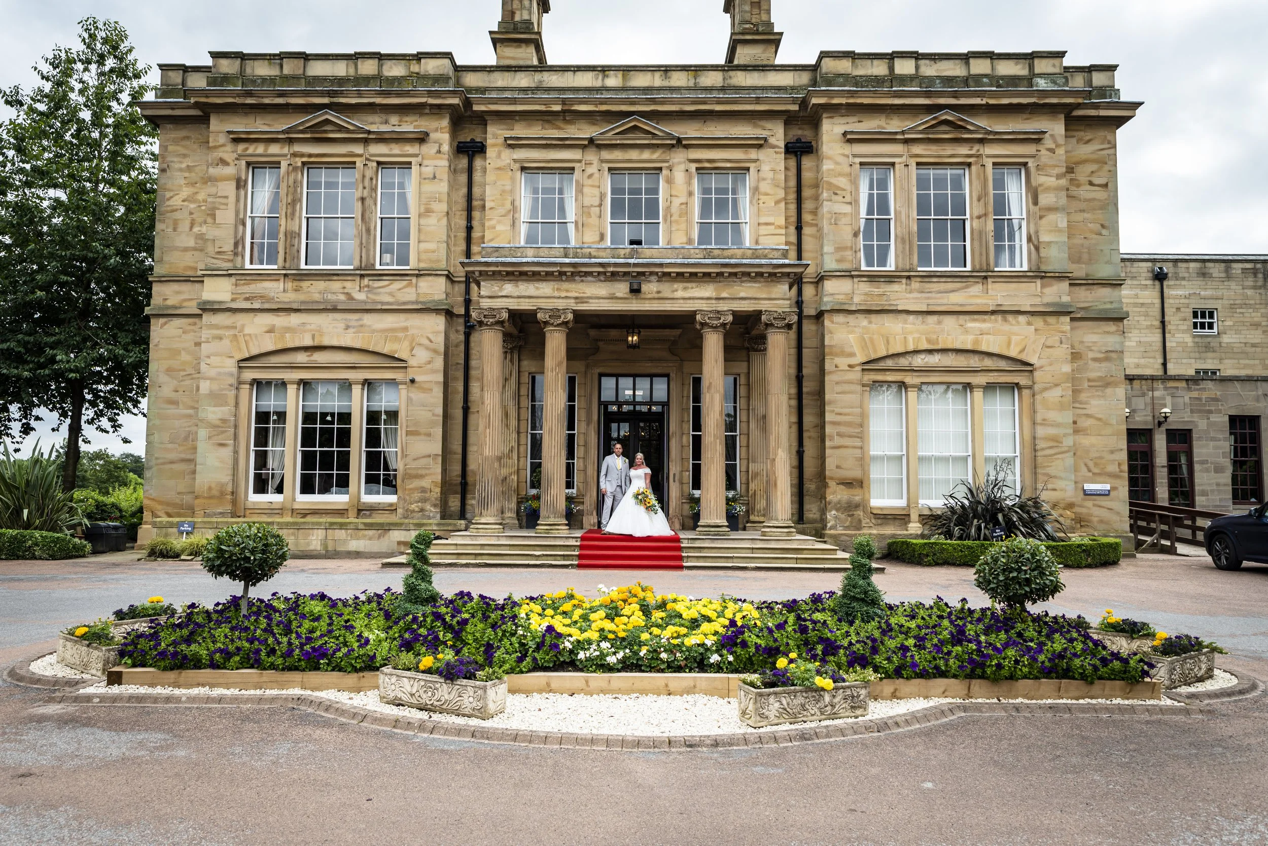 A bride and groom in wedding attire standing on a red carpet in front of a large, elegant, historic stone building with columns and tall windows. The building has a classical architectural style, with surrounded by well-maintained flower beds and gre