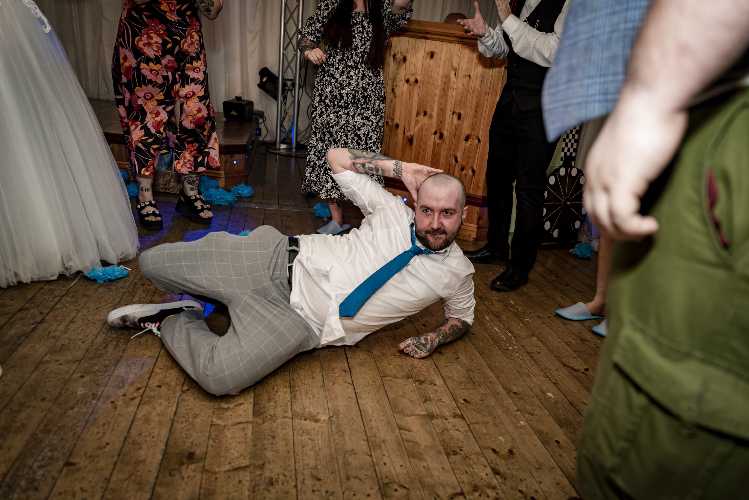 A man with tattoos and a beard lying on the wooden floor in a tuxedo shirt, plaid pants, and sneakers, holding his head with his hand. Several people are standing around him at what appears to be a party or celebration.