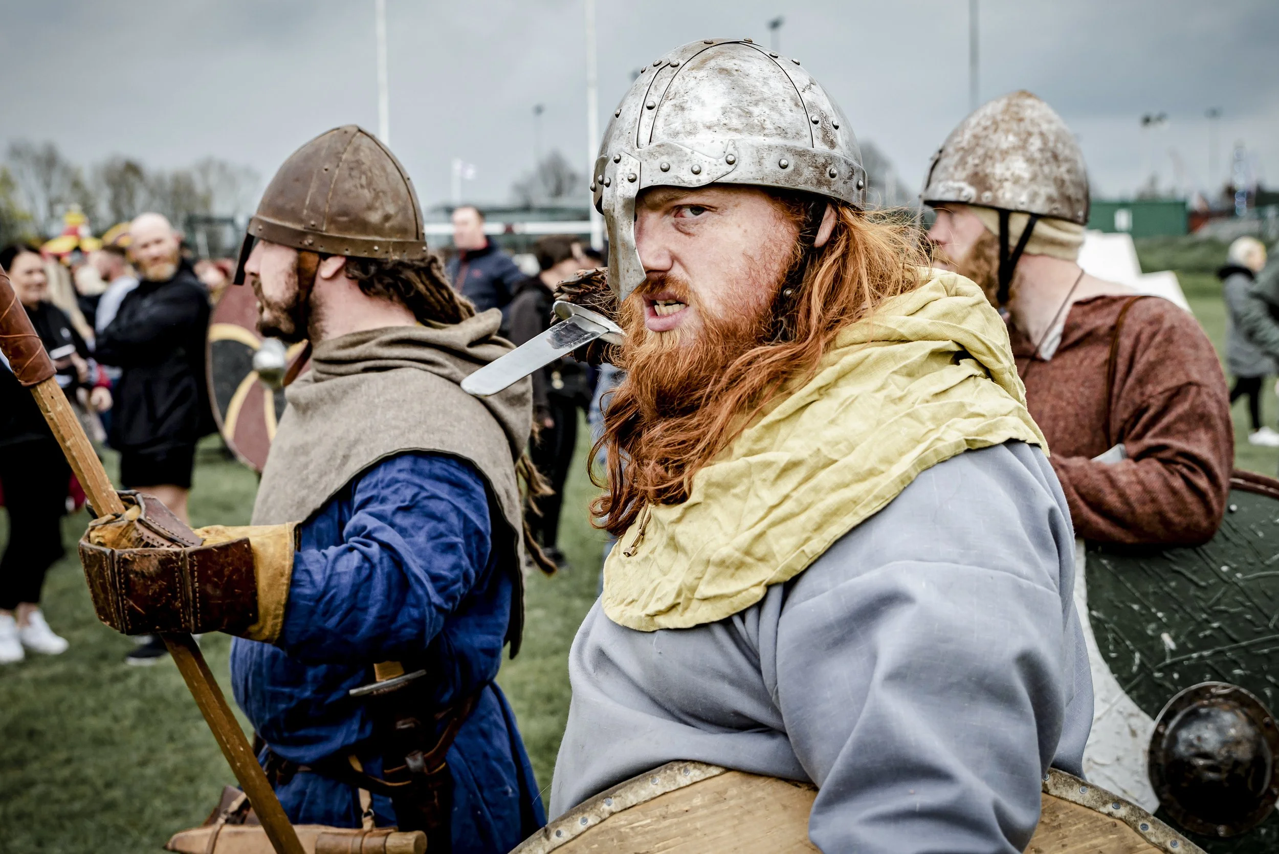 A group of people dressed as medieval warriors, wearing helmets and period costumes, at a historical reenactment event outdoors on a cloudy day.