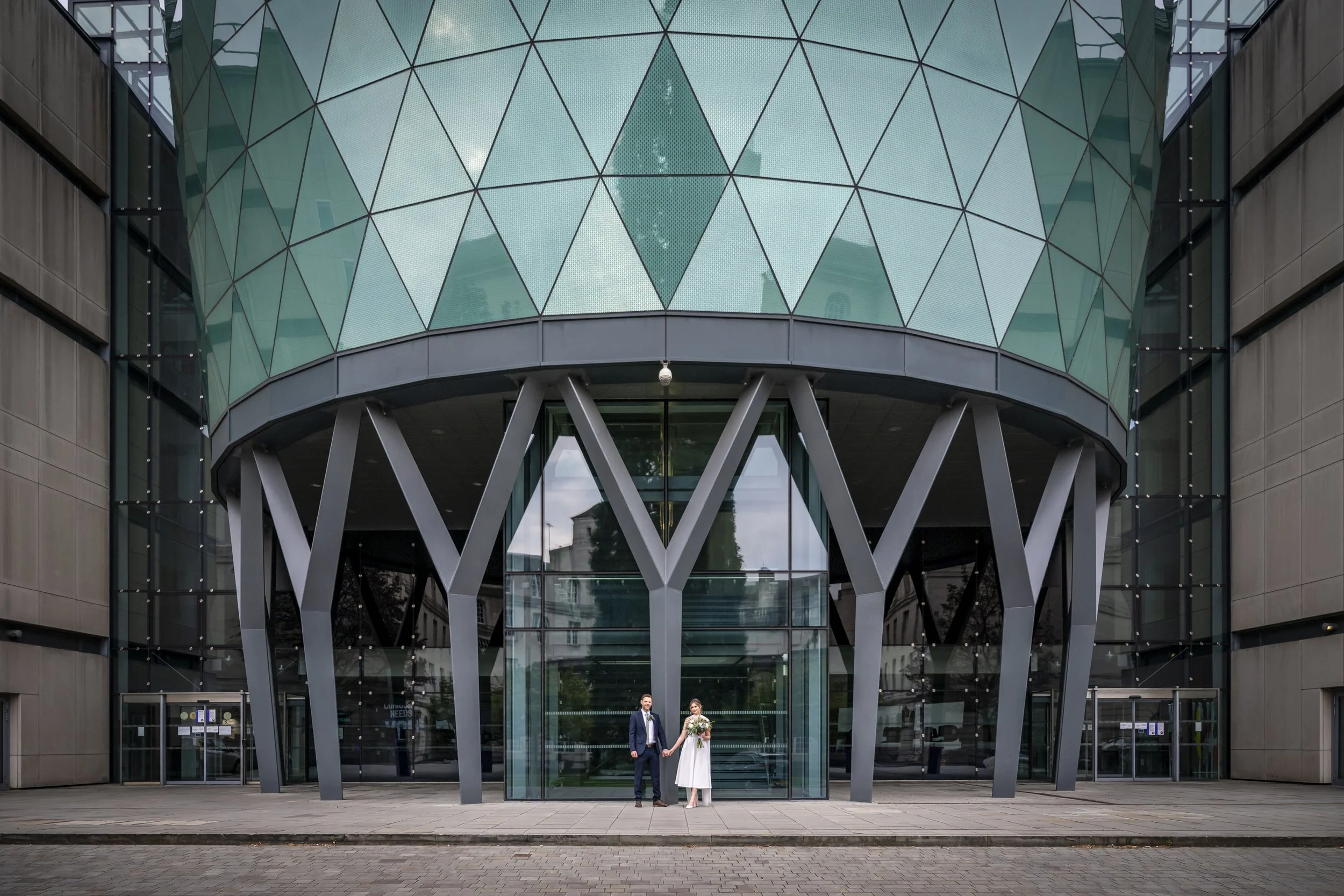 A bride and groom standing hand in hand outside a modern glass building with architectural support beams.