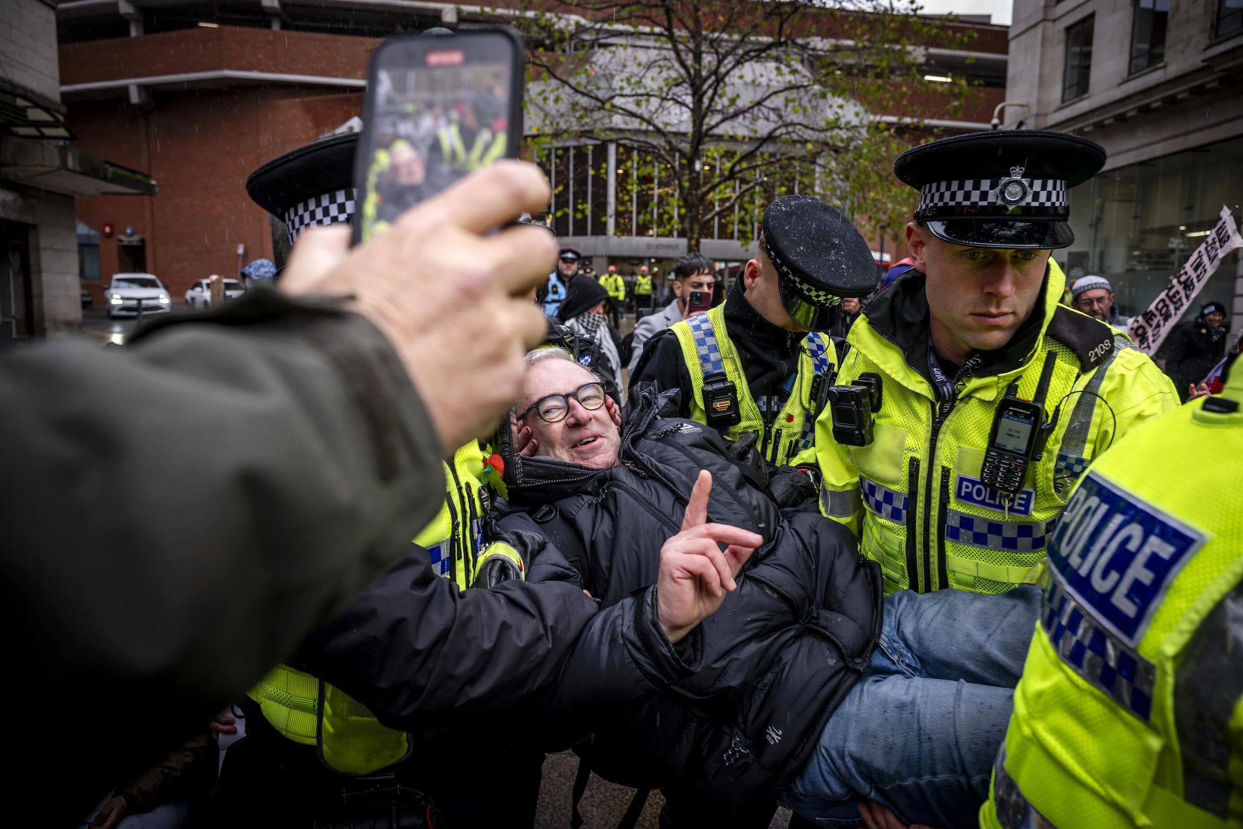 Police officers in high-visibility jackets arrest a man in a black puffer jacket during a protest, with people and buildings in the background. One person in the foreground is taking a photo or video with a mobile phone.
