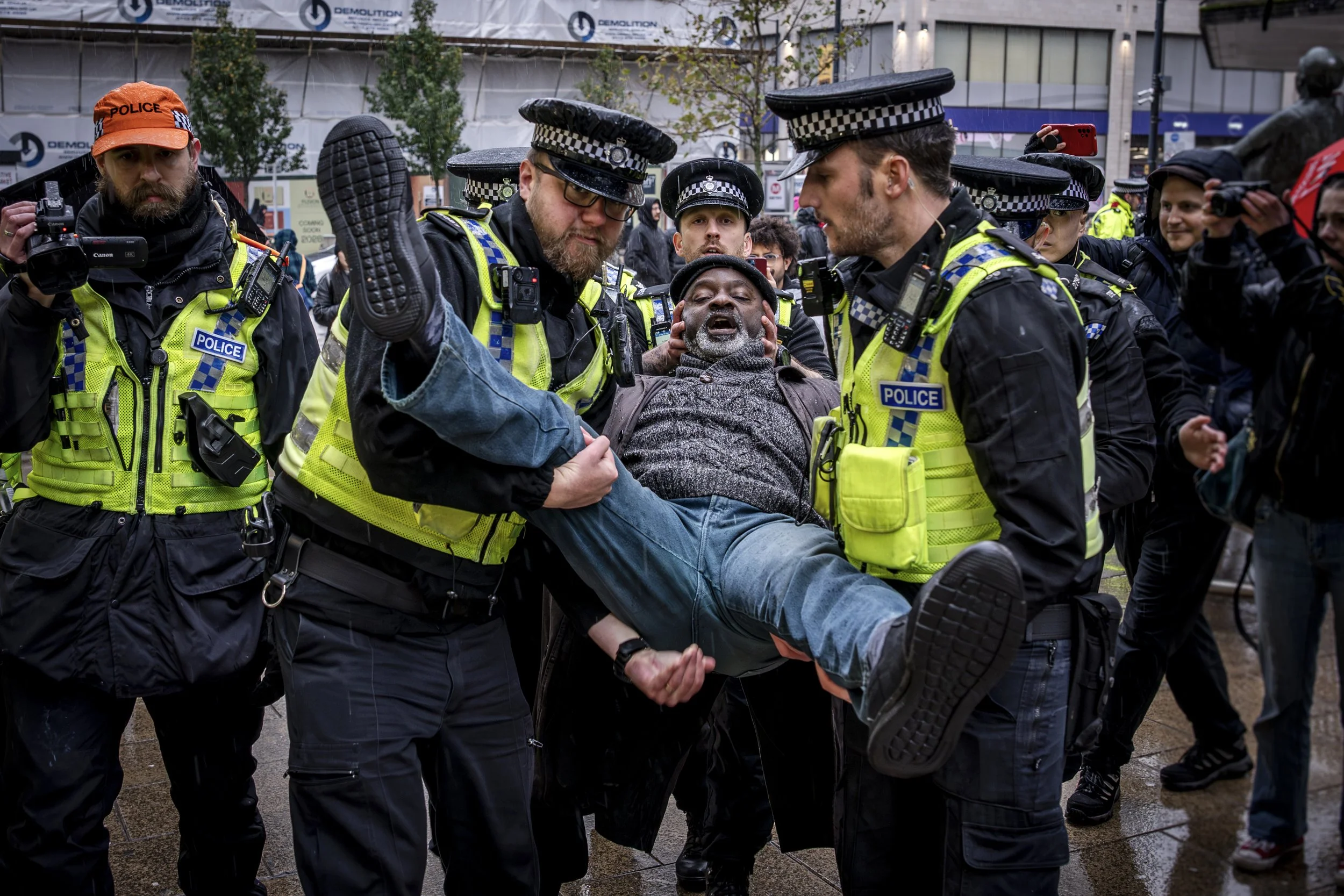 A group of police officers holding a man during a protest or demonstration, with some officers appearing to detain or assist him. Several onlookers and photographers are present in the background.