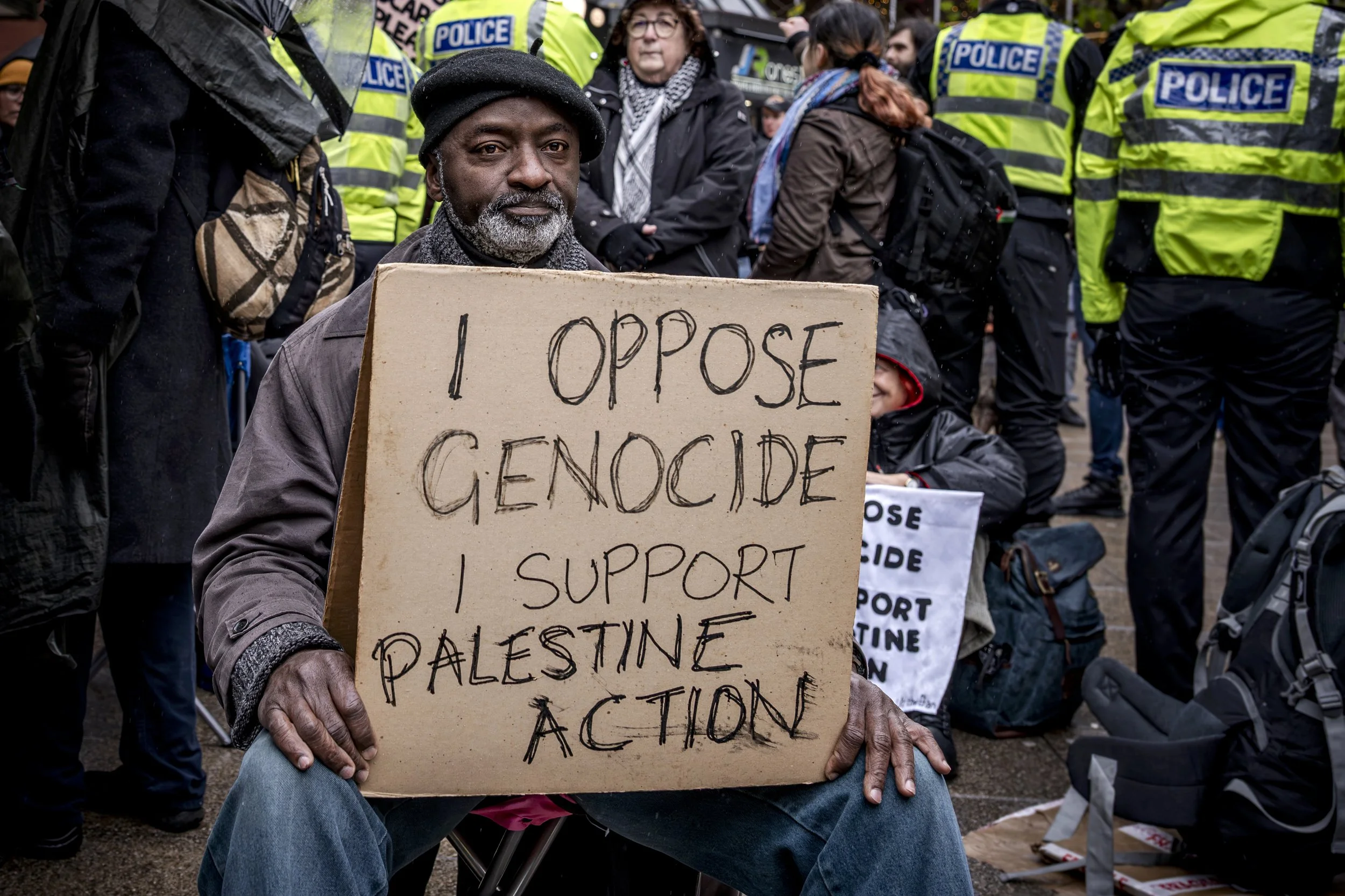 A man sitting and holding a cardboard sign that reads 'I oppose genocide, I support Palestine action' during a protest or rally, with police officers and other protesters in the background.