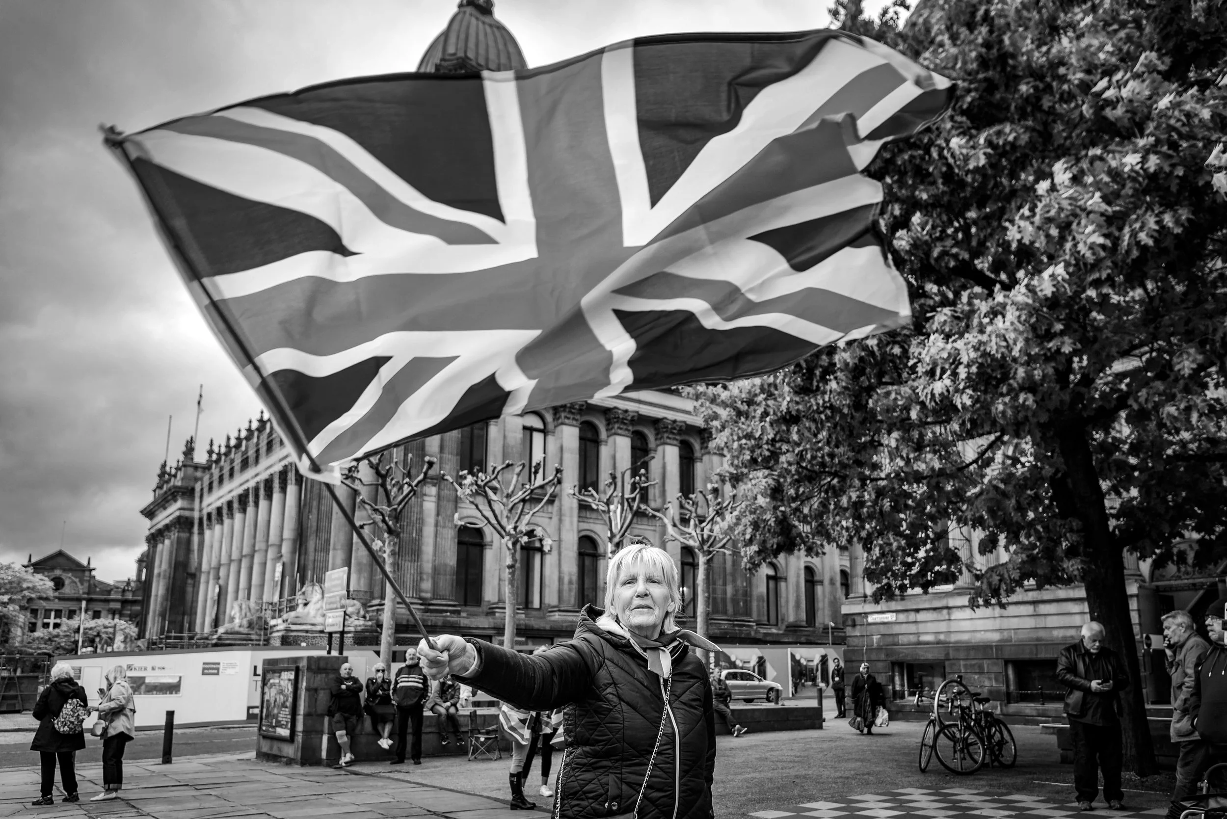 A woman holding a Union Jack flag in a cityscape with historic buildings and several people in the background.