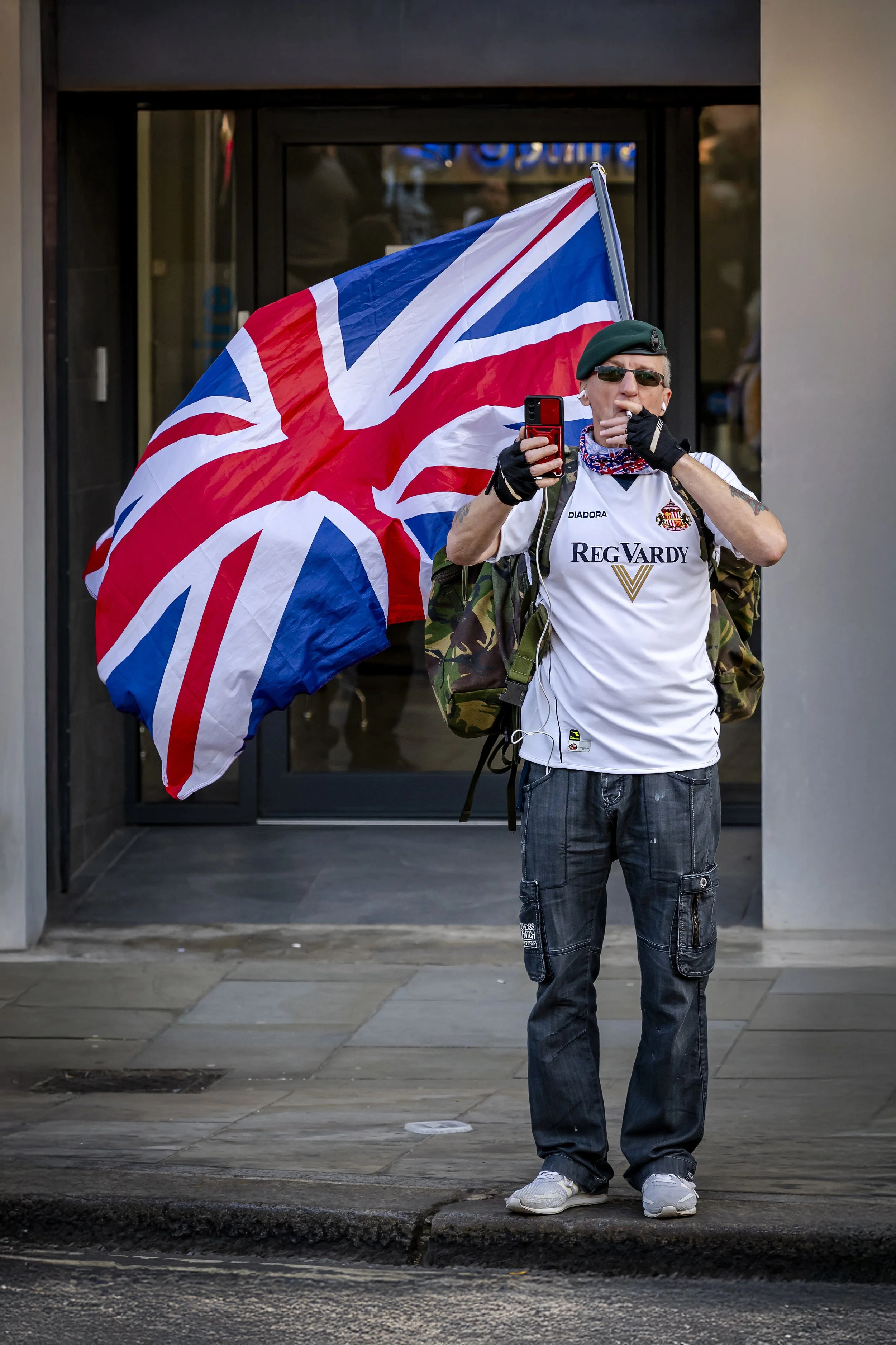 A man standing on a city sidewalk holding a large British flag. He is wearing a green beret, sunglasses, a white sports jersey, and camouflage backpack. He's making a smoking gesture with his left hand while holding a red phone with his right. The ba
