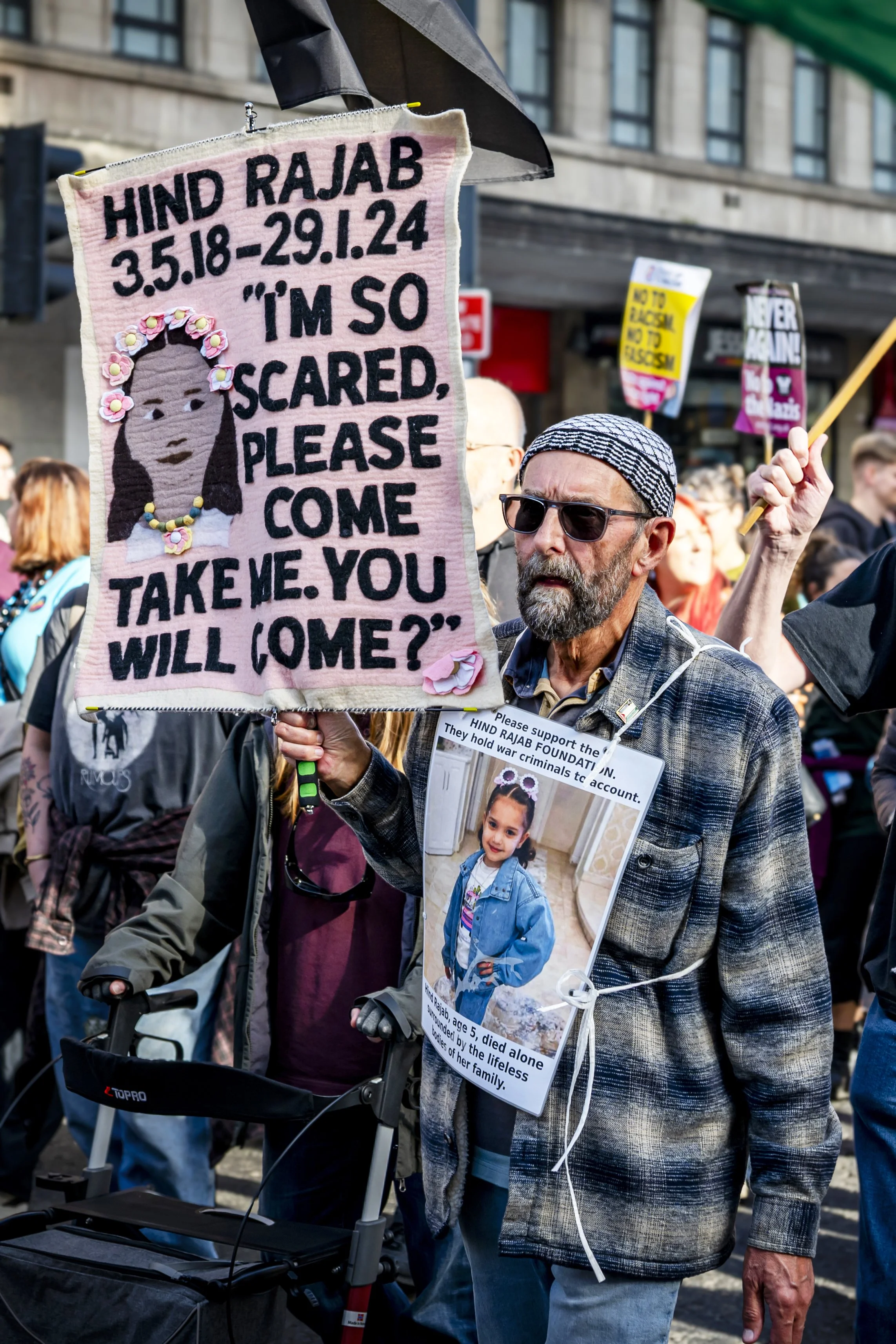 A man at a protest holding a handmade sign featuring a girl’s portrait, with text about fearing the Hind Rojab Foundation and a plea for help, surrounded by other protesters holding signs.