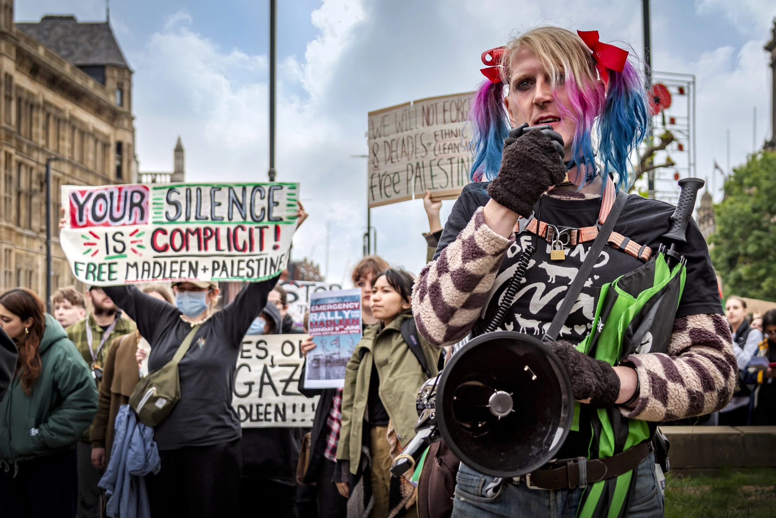 A woman with colorful hair holding a megaphone at a protest, surrounded by people holding signs supporting Palestine and speaking out against silence and injustice.