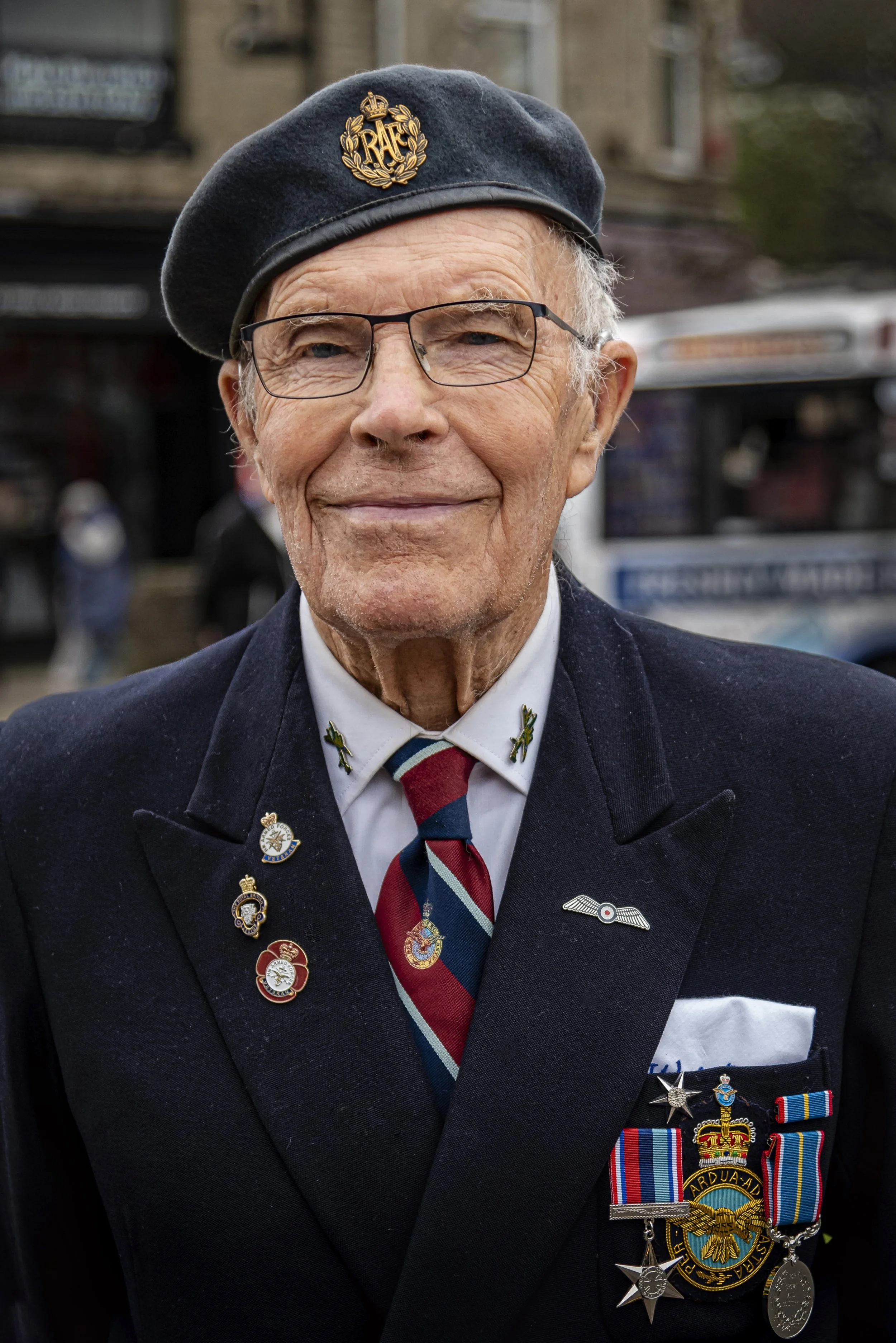 An elderly man wearing a navy parade uniform with medals and pins, a tie, glasses, and a navy beret with a gold emblem, smiling on a city street.