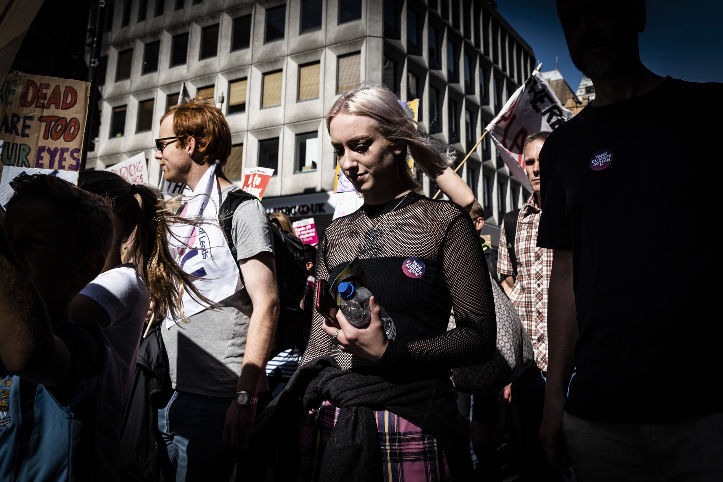 Group of people participating in a street protest or rally, holding signs and flags, in front of a modern building on a sunny day.