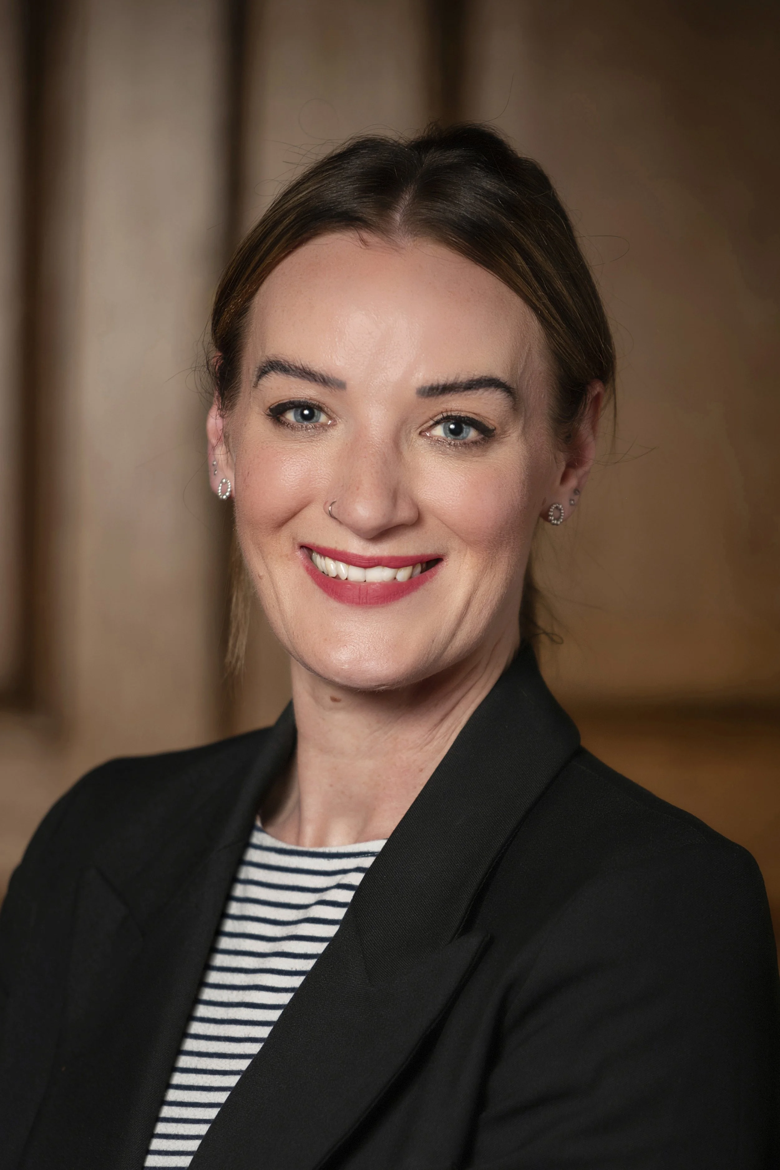 Professional woman with short brown hair, blue eyes, and light skin, smiling, wearing a black blazer over a striped shirt, with earrings and a nose piercing. The background is blurred.