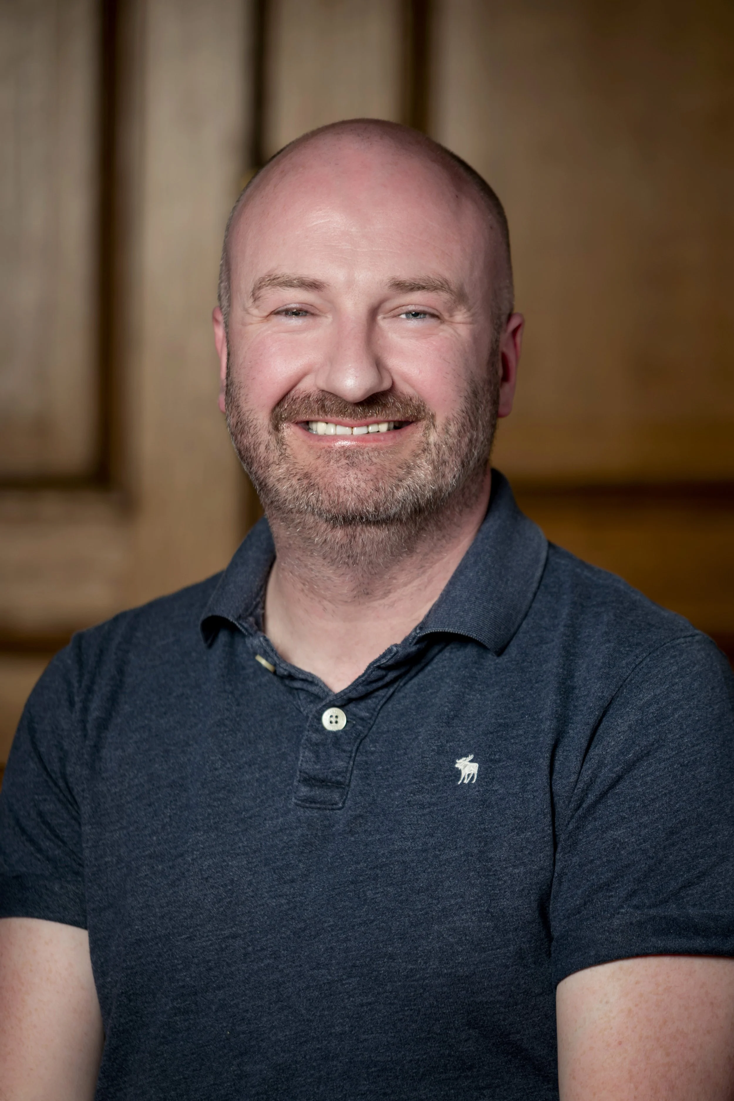 Headshot of a smiling, bald man with a beard, wearing a navy blue polo shirt, in front of a wooden background.