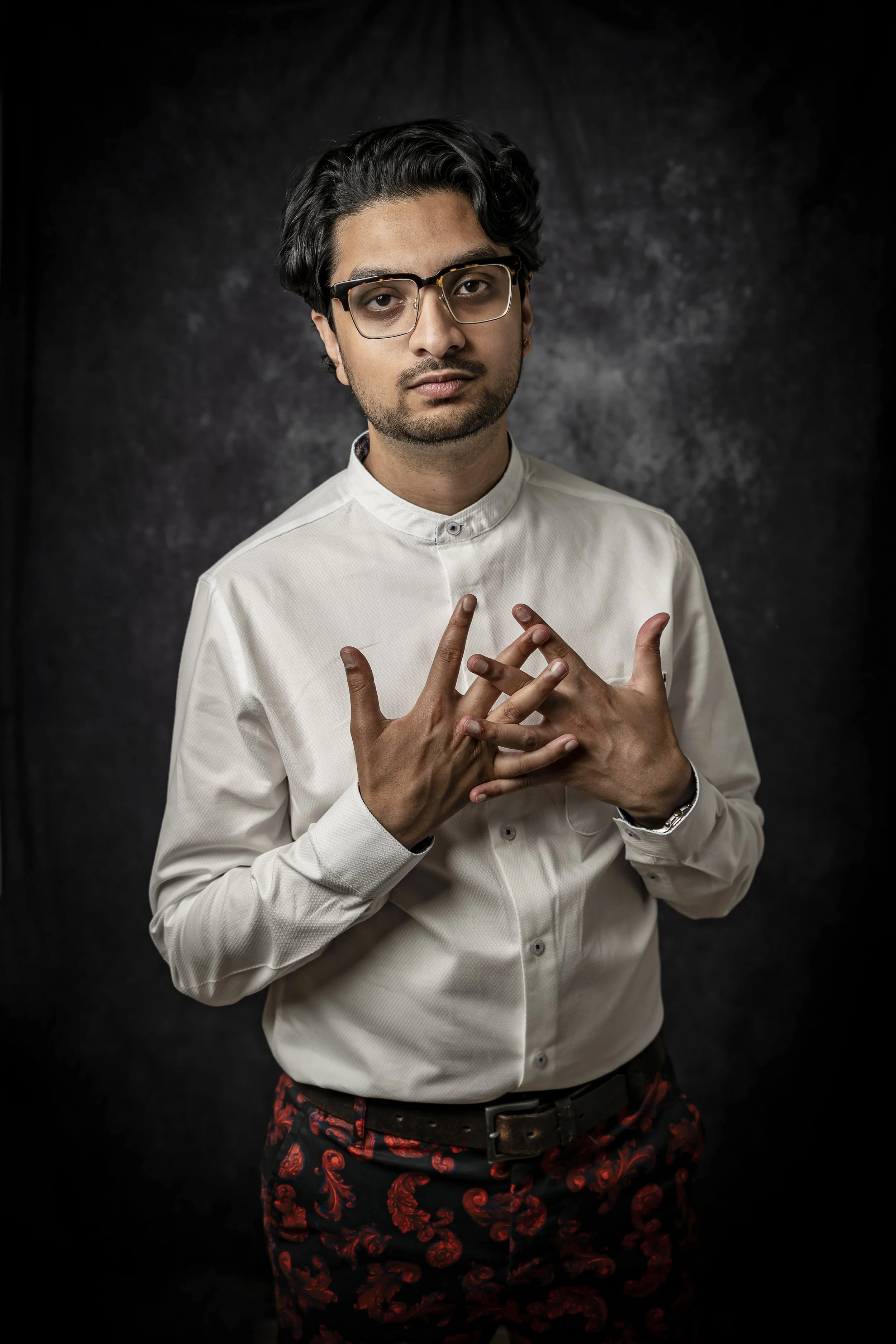 A young man with glasses, dark hair, and a light complexion posing against a dark, textured background. He is wearing a white button-up shirt and dark pants with a red floral pattern, with his hands gently clasped together at chest level.