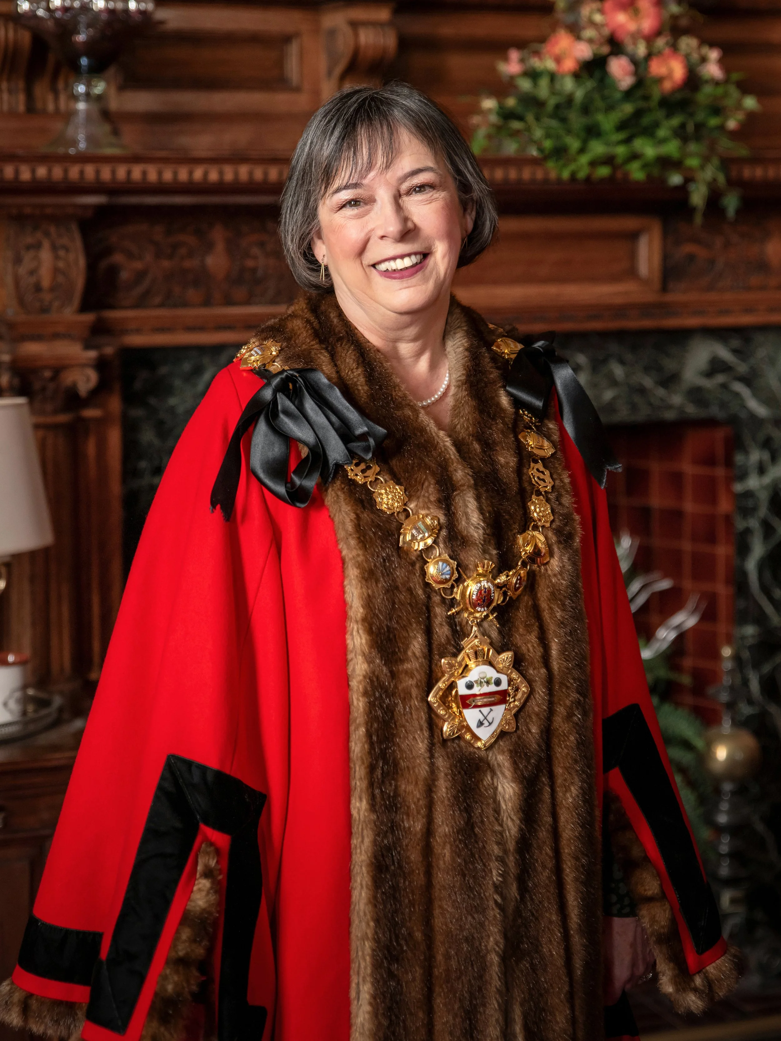A woman wearing a red ceremonial robe with black accents, a fur stole, a gold ceremonial chain with a crest, standing in a richly decorated room with wooden furniture and a floral arrangement in the background.